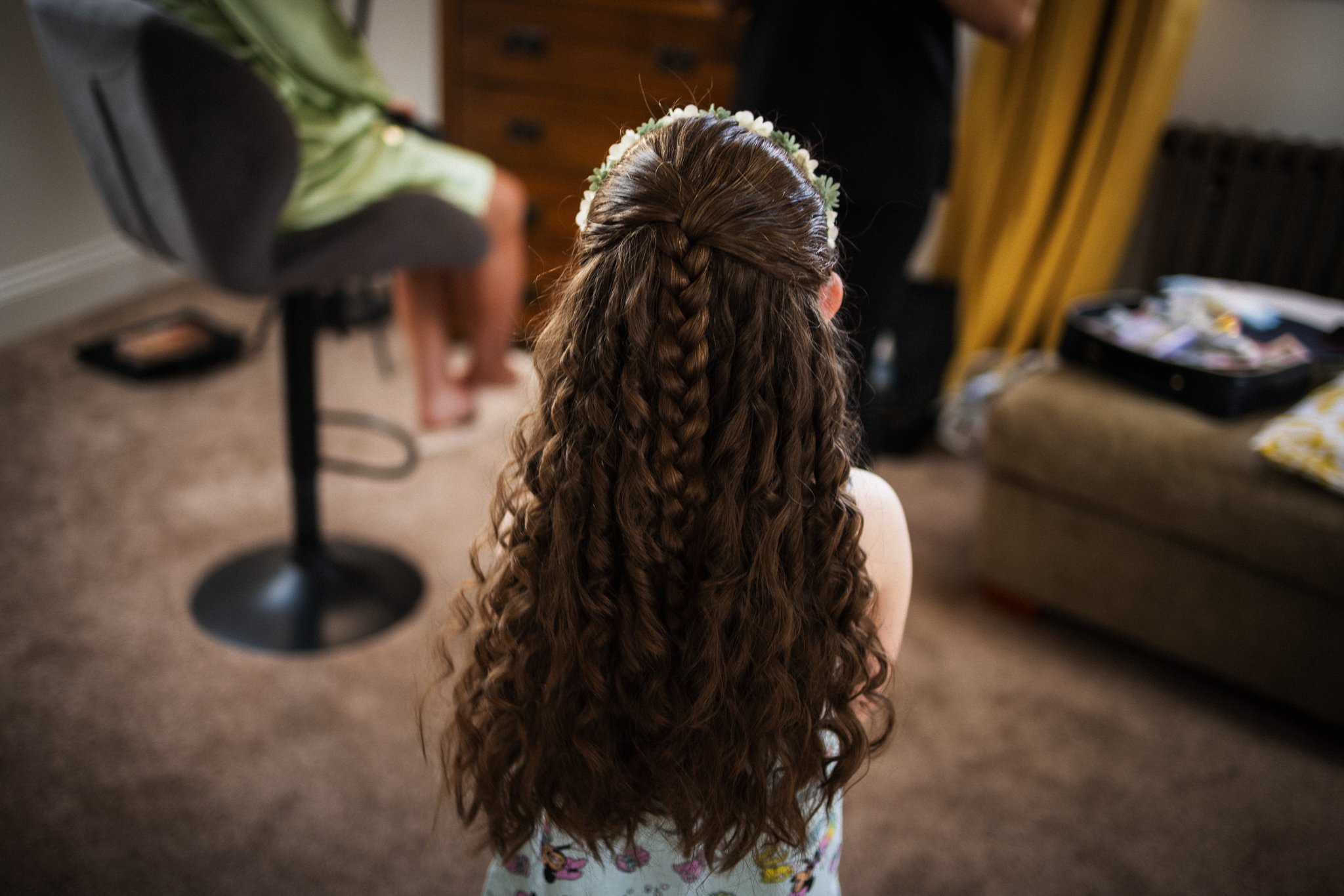 Back view of a girl with long, curly hair wearing a floral headband, sitting on the floor, with a dresser and a person blurred in the background.