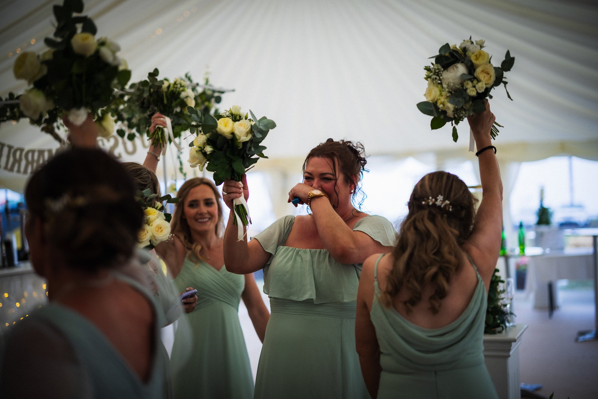 Women in matching light blue dresses holding bouquets at a celebration, possibly a wedding reception, inside a decorated event tent.