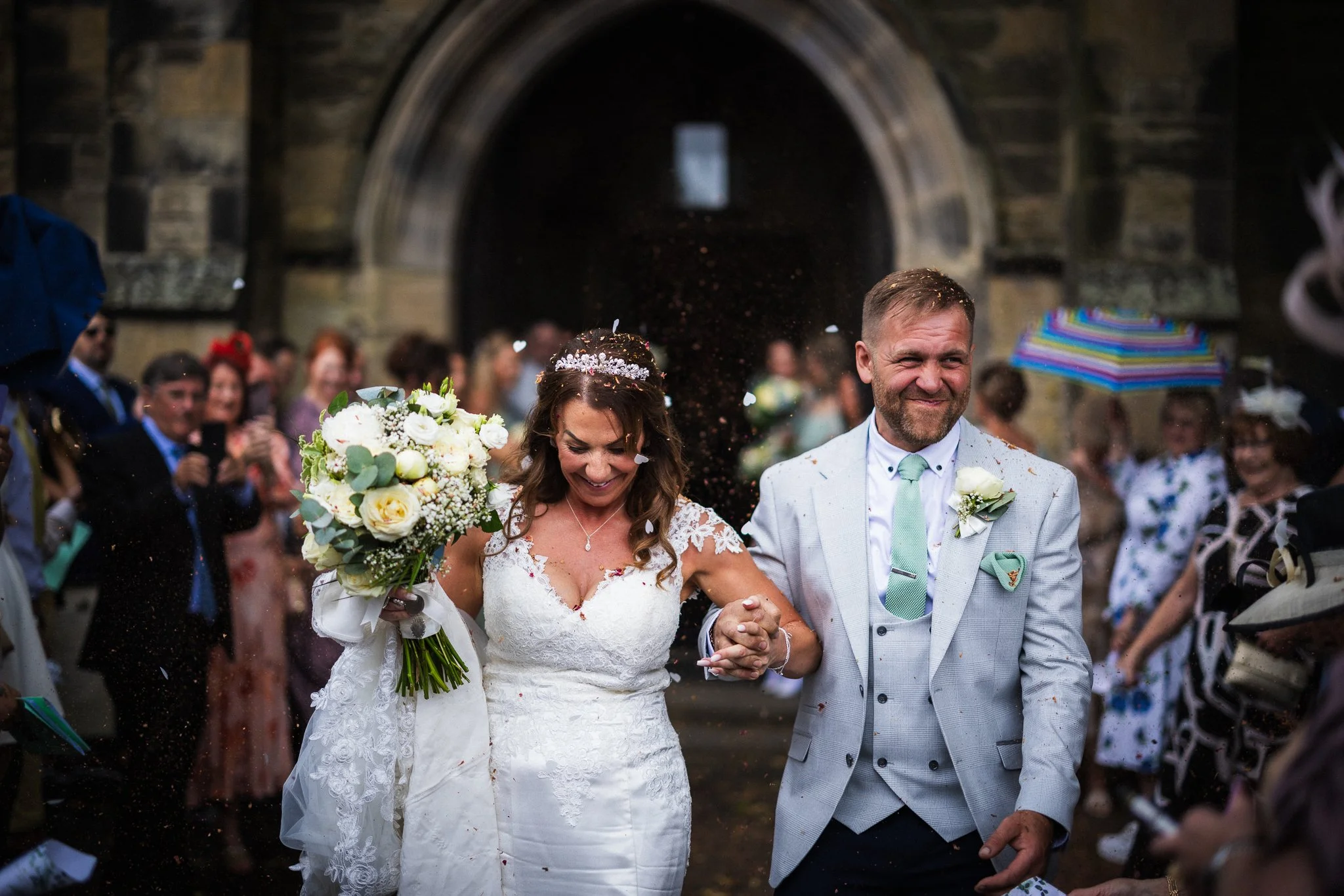 A bride and groom walking hand in hand at their wedding, surrounded by guests, with confetti falling around them outside a stone building.