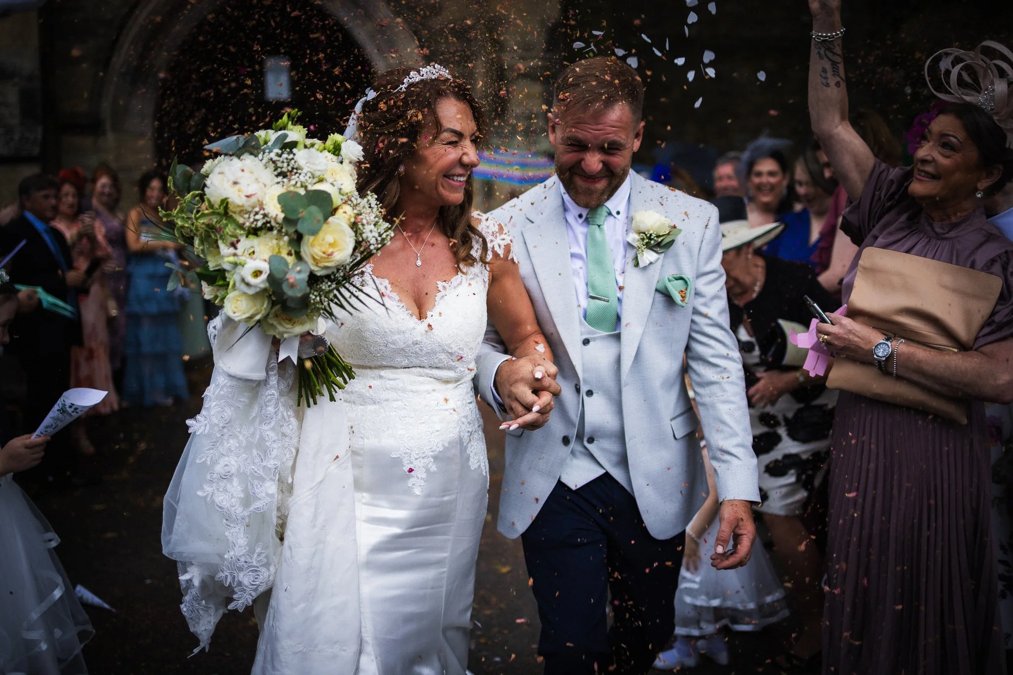 A bride and groom smiling and holding hands during their wedding celebration, surrounded by guests throwing confetti.