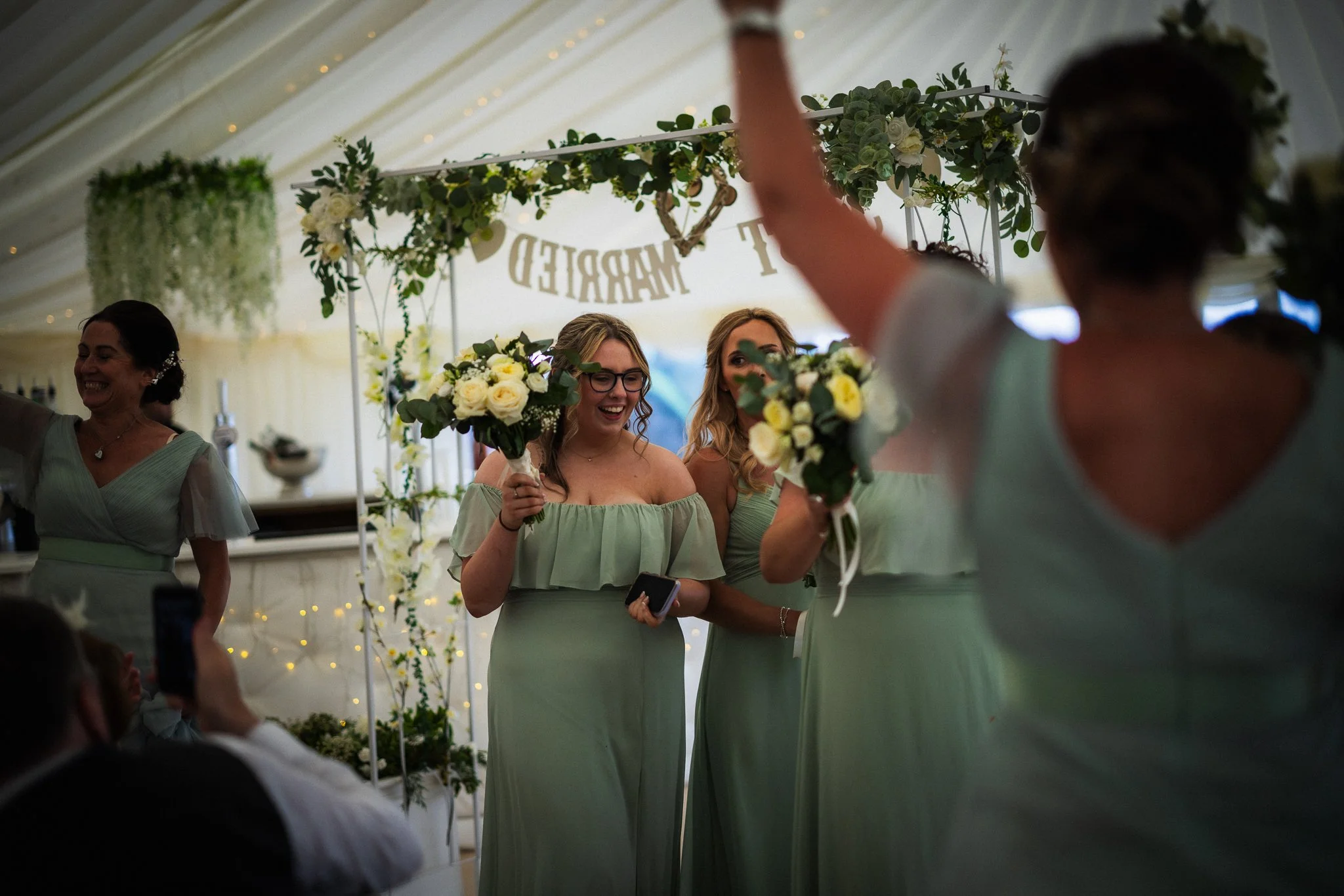 Women in light green dresses holding bouquets during a celebration or wedding reception, with a floral and greenery decorated backdrop.