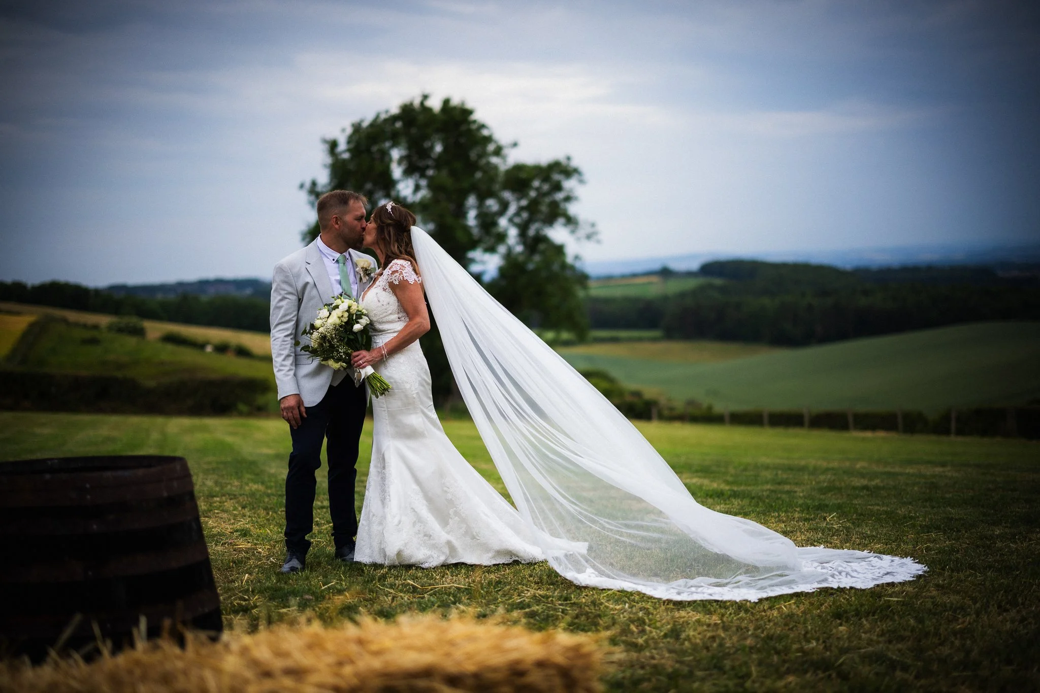 A bride and groom kiss outdoors on a cloudy day, with a large tree and rolling fields in the background. The bride is wearing a white wedding dress with a long train and a veil, holding a bouquet of white roses, and the groom is dressed in a light gr