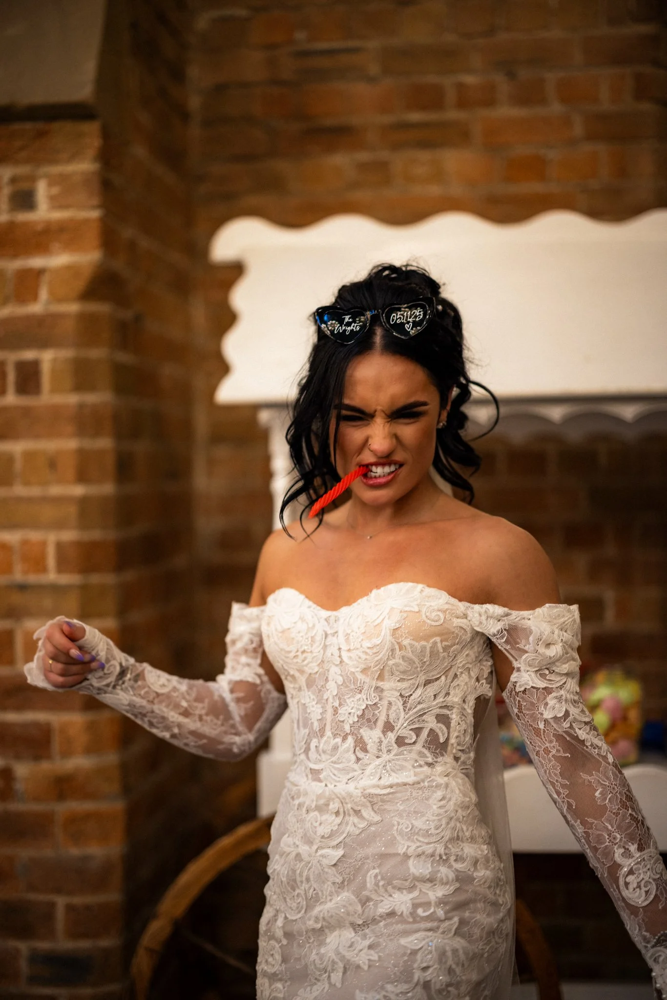 A woman in a lace off-shoulder wedding dress making a grimace while holding her arm out with a clenched fist. She has dark hair with sunglasses on her head and is chewing a piece of red string.