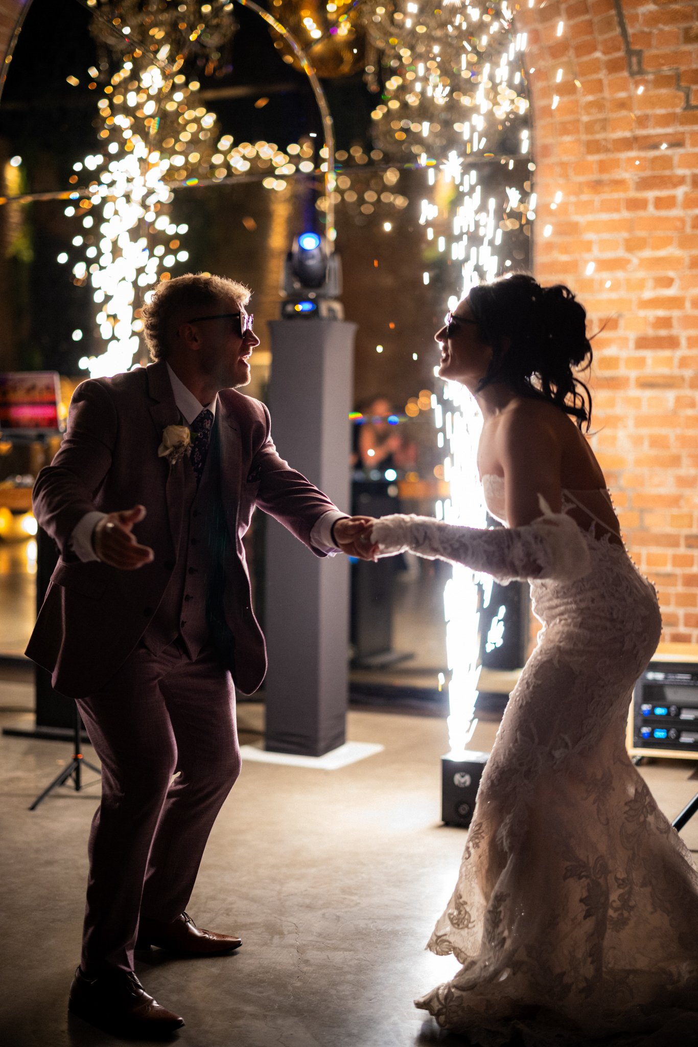 A man and woman in formal attire dancing at night, with sparklers and a brick wall in the background.
