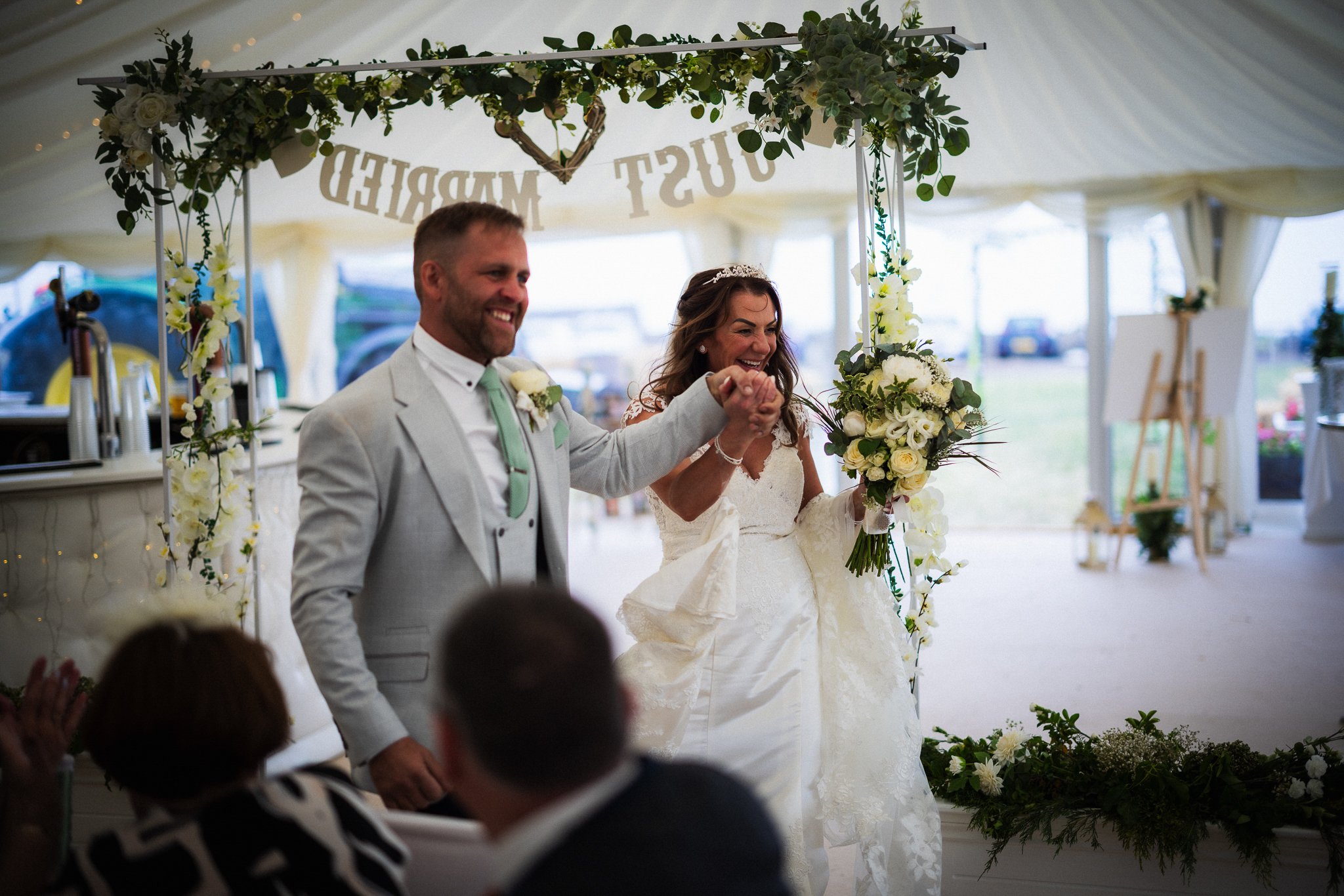 A happy bride and groom at their wedding reception, holding hands, standing under a floral arch with a "Just Married" banner, surrounded by guests and floral decorations in a tented outdoor setting.
