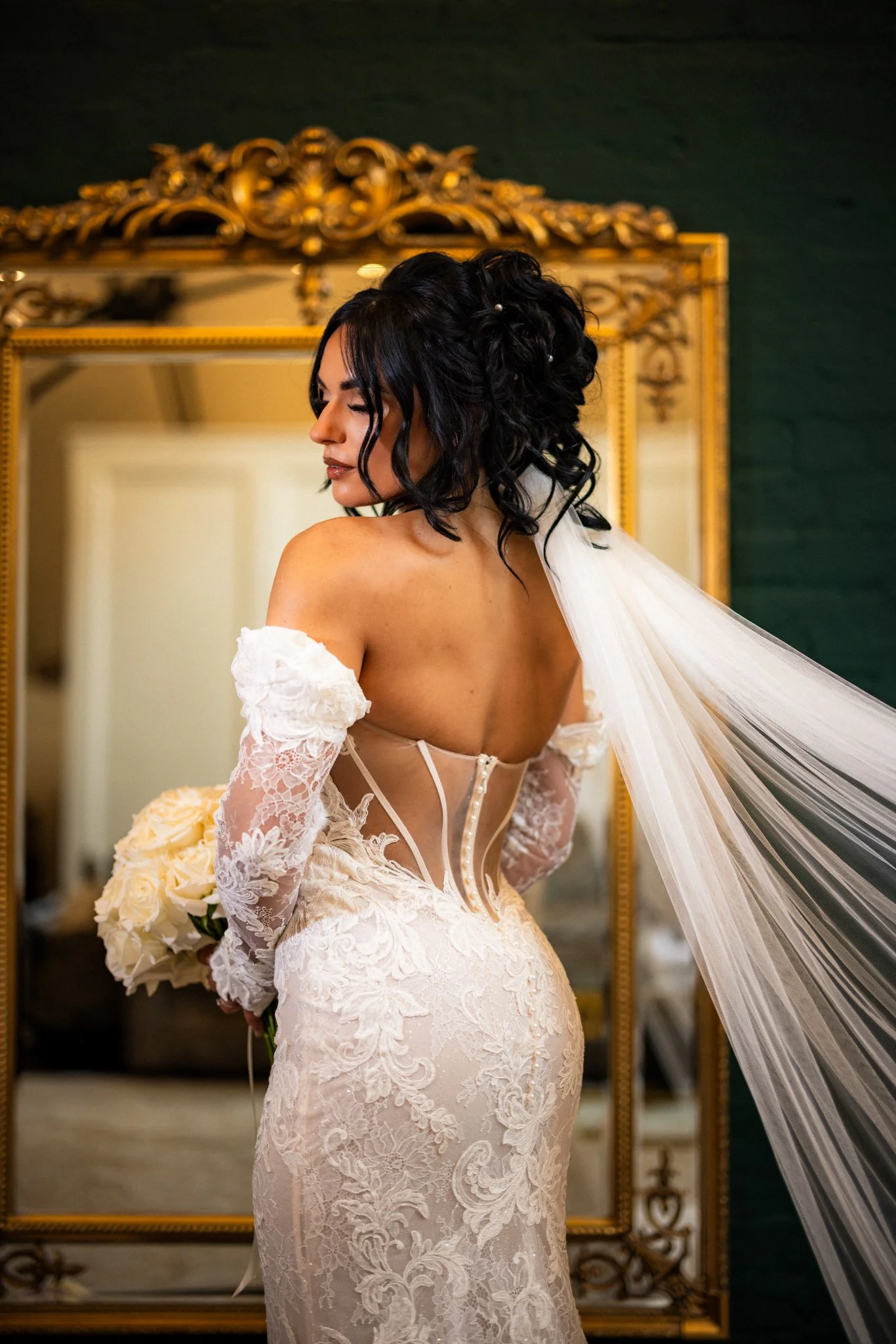 A woman with dark, curly, styled hair and a pearl hairpin, wearing a white lace wedding dress with off-the-shoulder lace sleeves and a sheer back, stands in front of a gold ornate mirror, holding a bouquet of white roses.