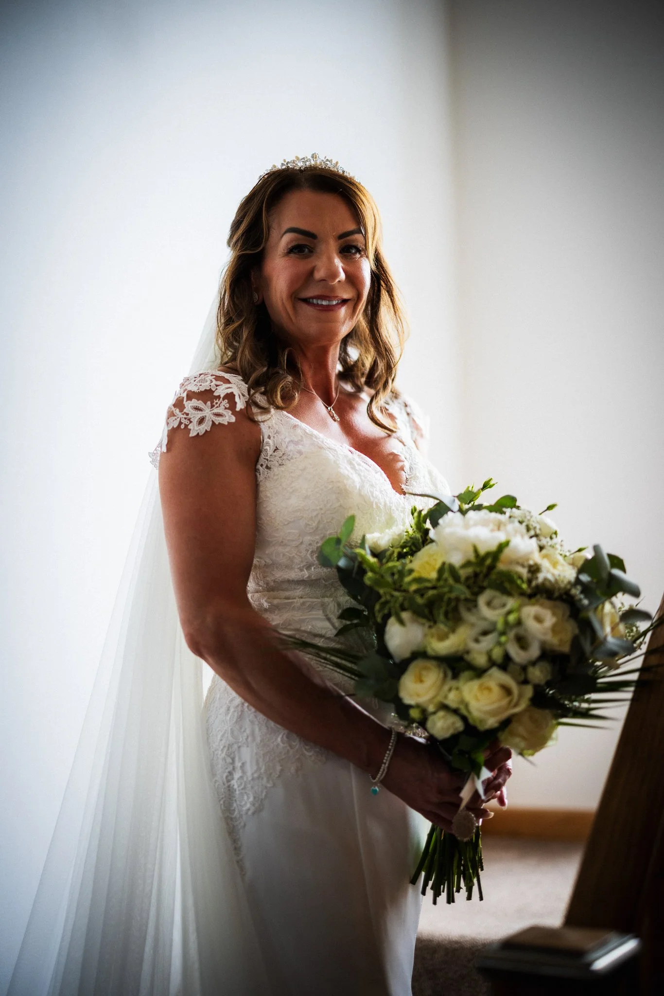 A bride wearing a white lace wedding gown with floral embroidery on the sleeves, holding a bouquet of white and cream flowers, standing indoors near a plain white wall, smiling at the camera.