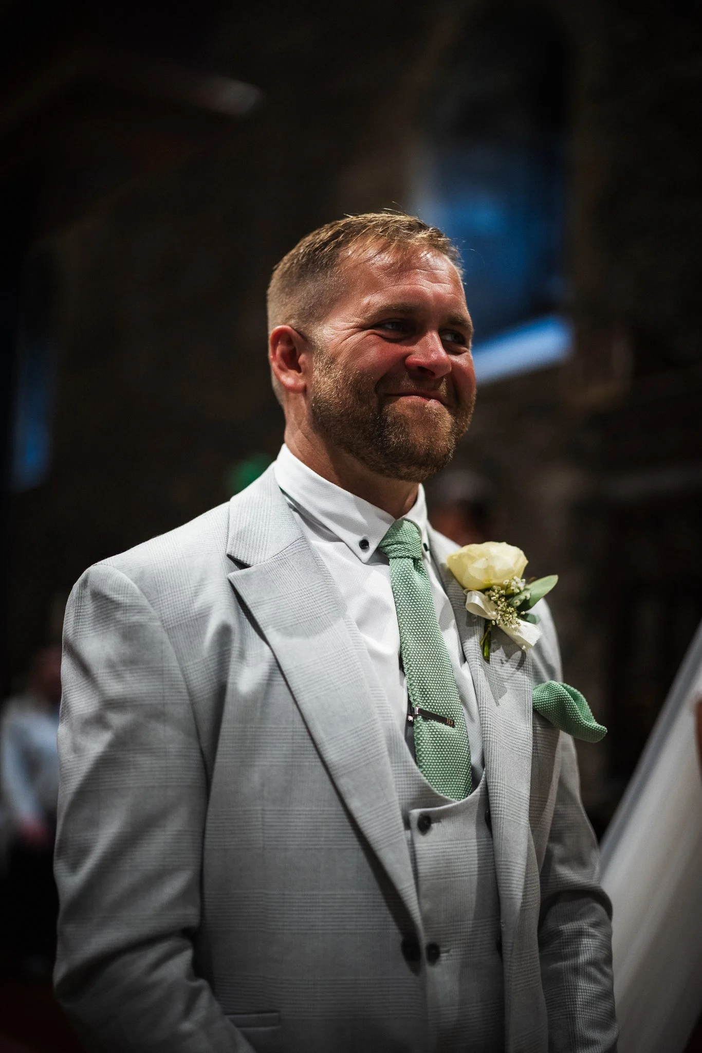 A man in a light grey suit with a green tie and checkered pocket square, smiling and holding a white flower boutonniere.