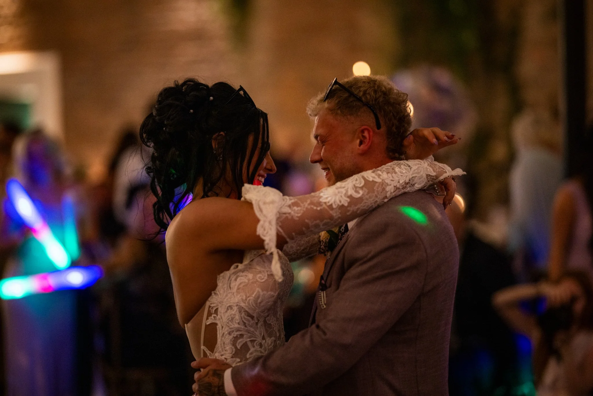 A couple dancing closely together at an indoor event during the evening. The woman is wearing a lace dress, and the man is in a suit. They are smiling and enjoying the moment with blurred colorful lights in the background.