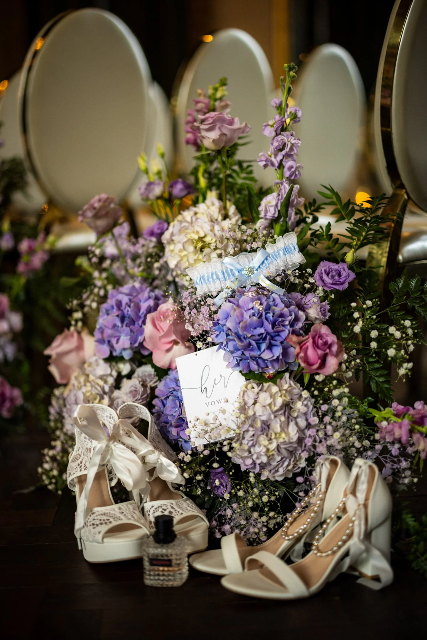 A collection of white and beige high-heeled shoes with decorative straps and pearls, arranged on a dark surface. Behind the shoes are bouquets of purple, pink, and white flowers, with a wedding card and a small bottle of perfume.