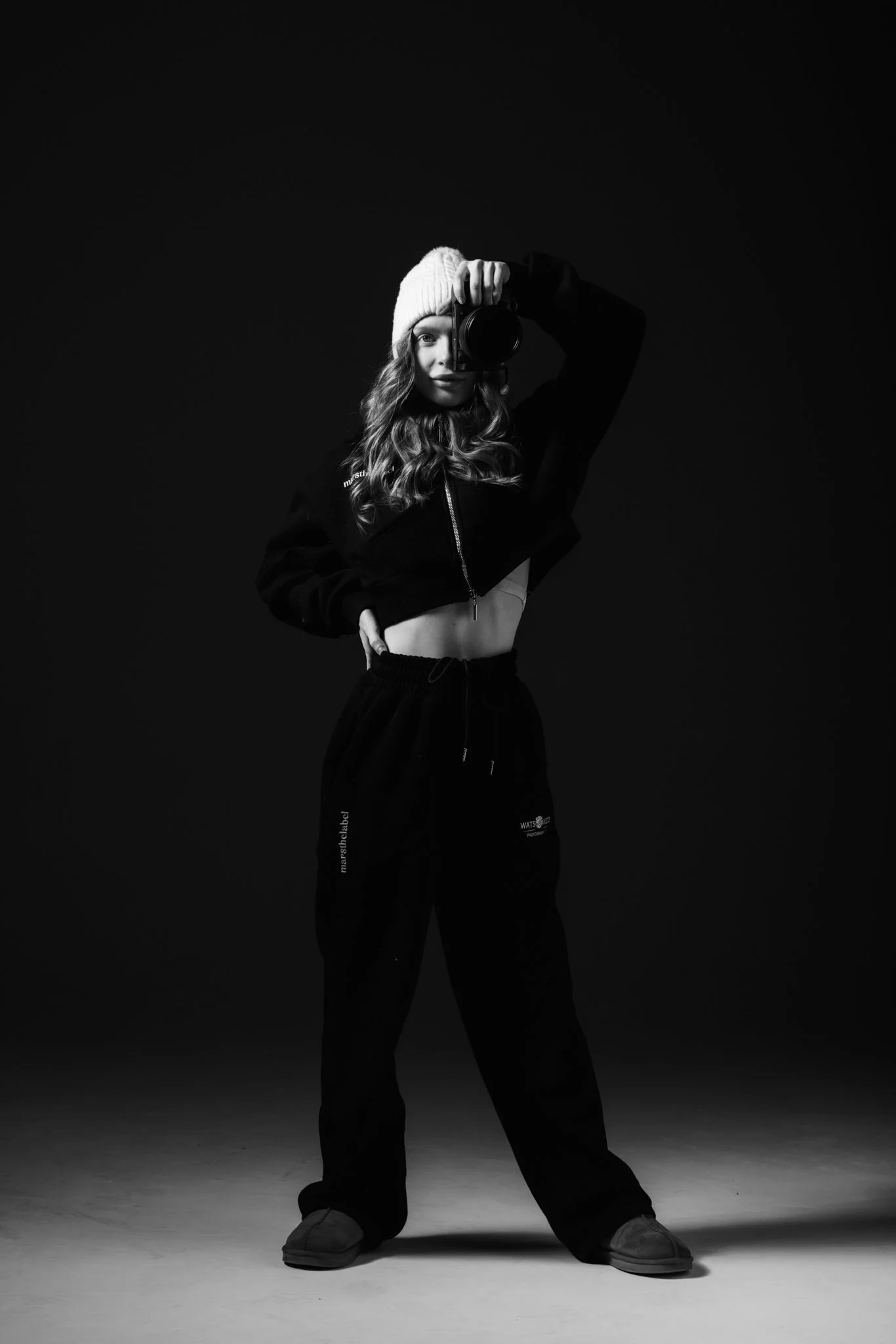 A young woman with long curly hair wearing a beanie, a cropped jacket, sweatpants, and sneakers, taking a self-portrait with a camera in a dark studio setting.