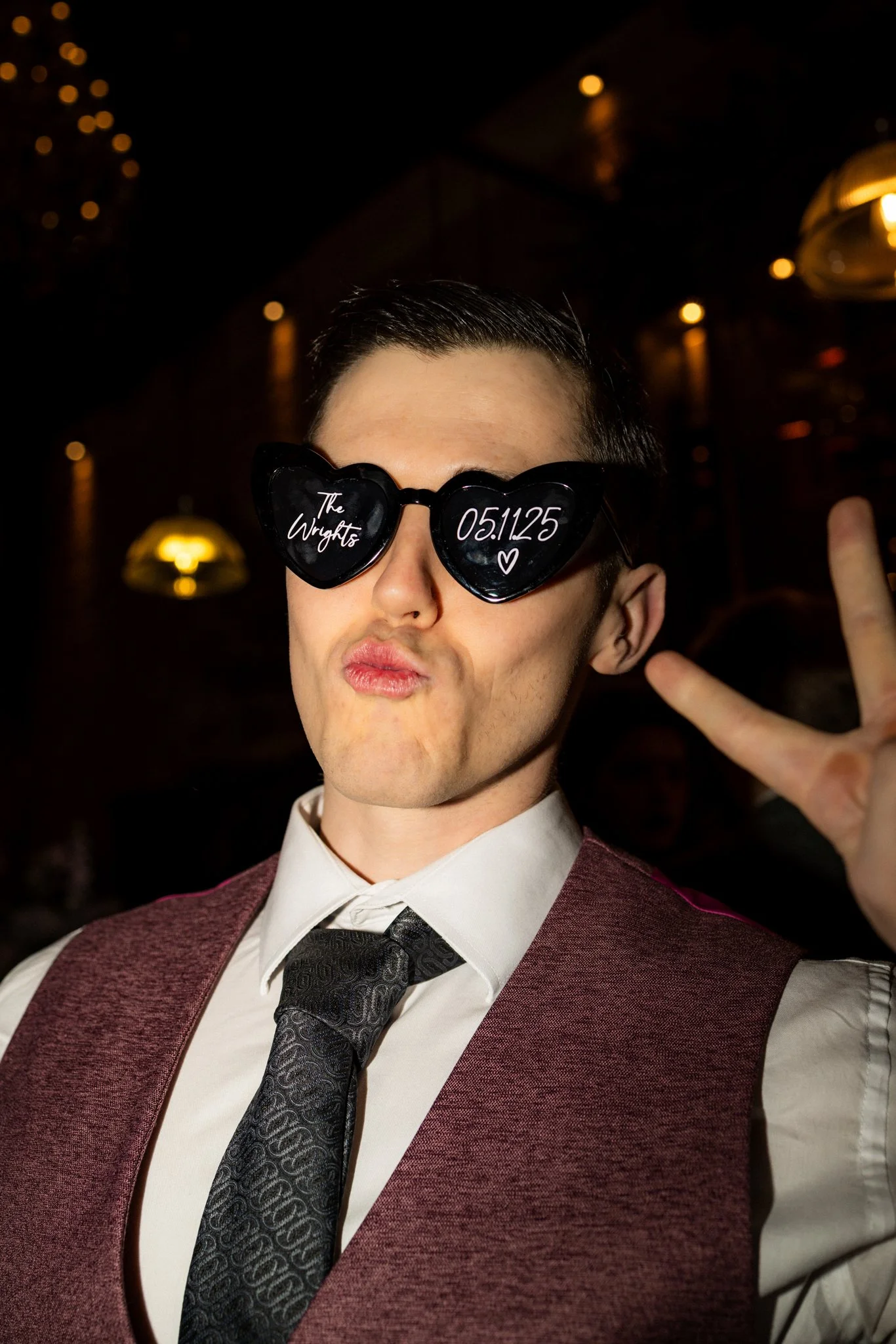 A person wearing heart-shaped sunglasses with writing, a white shirt, a dark tie, and a maroon vest in a dimly lit indoor setting.