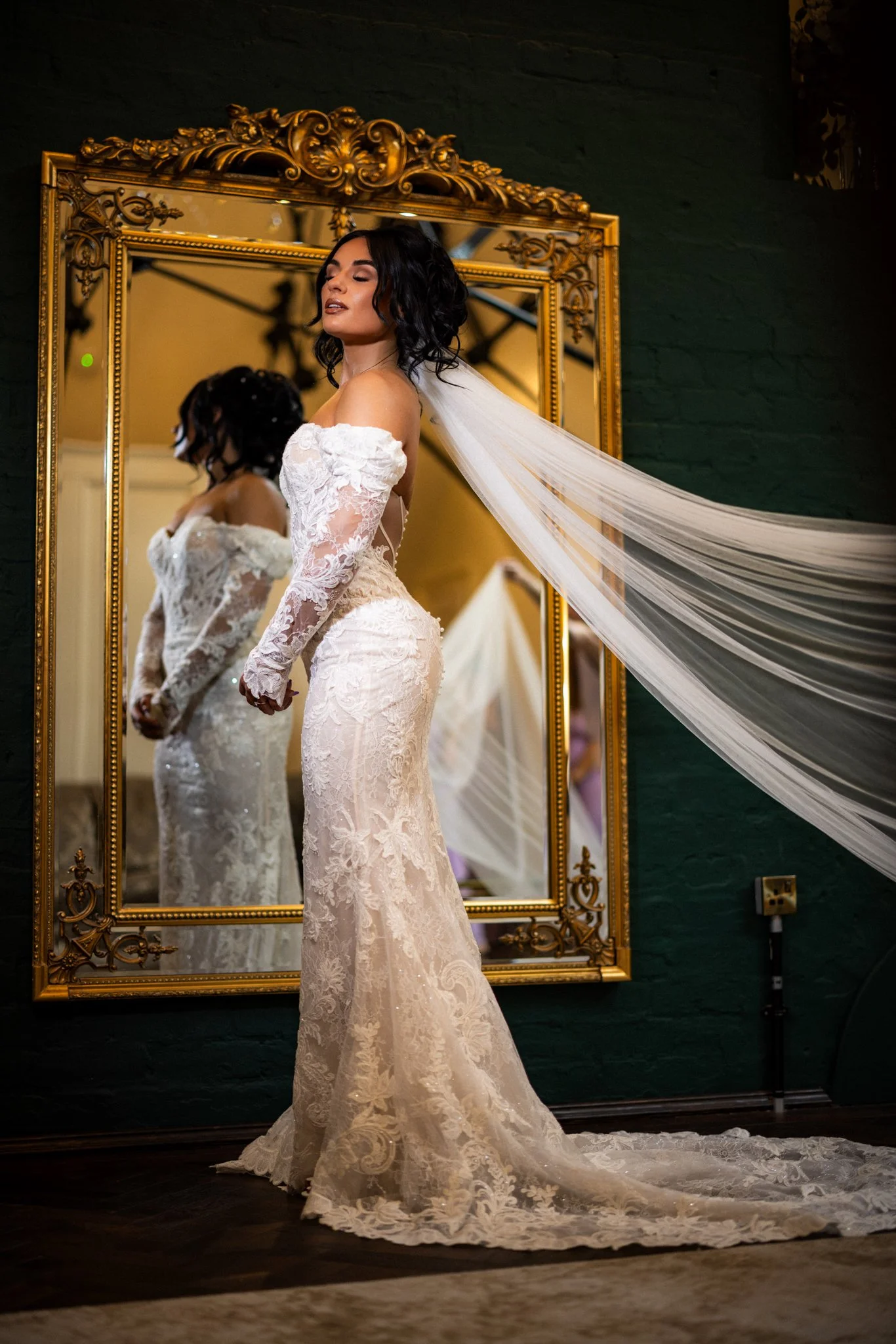 A woman in a white lace wedding dress stands in front of an ornate gold-framed mirror, adjusting her dress and veil.