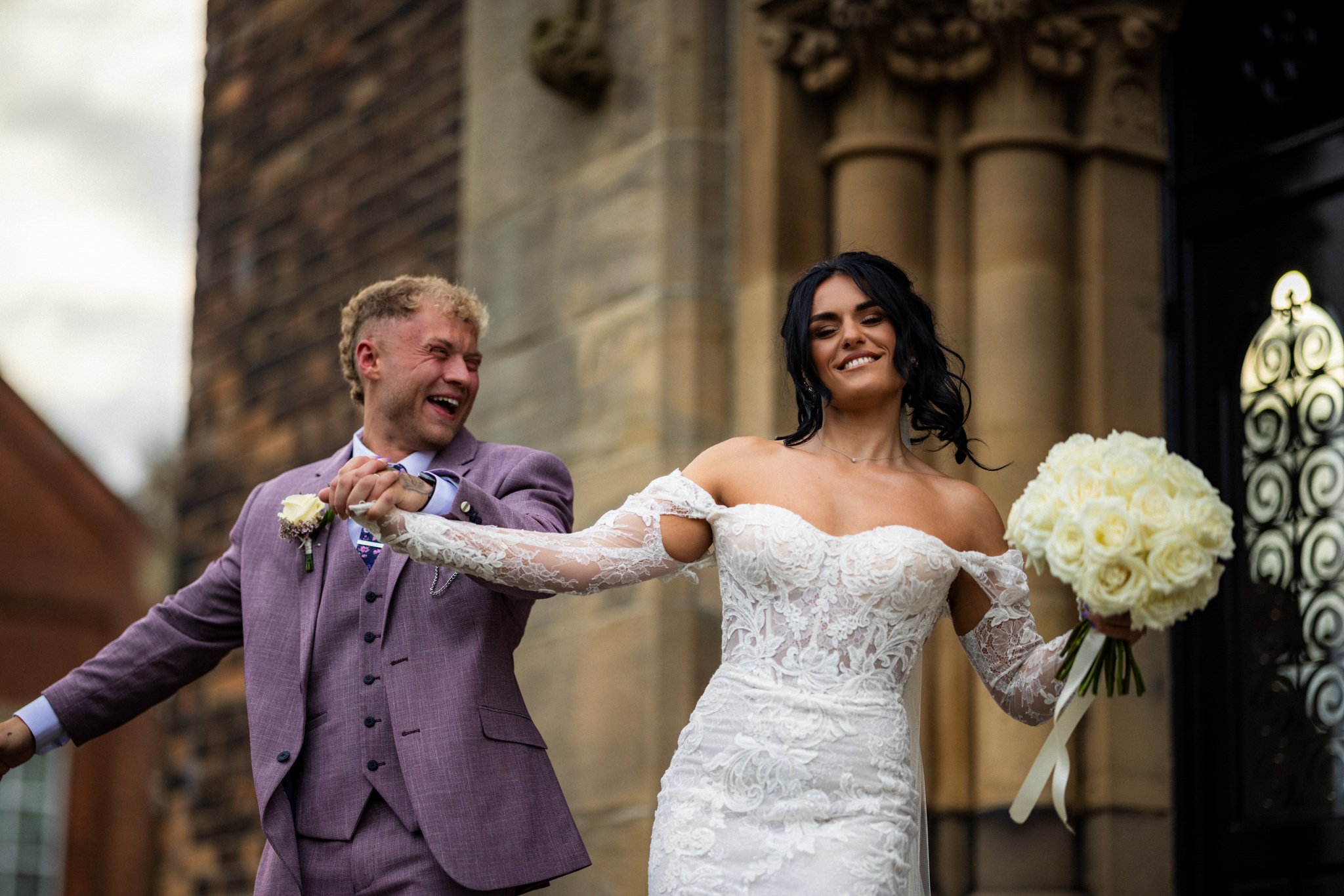 A bride and groom smiling and holding hands outside a church, with the bride holding a bouquet of white roses.