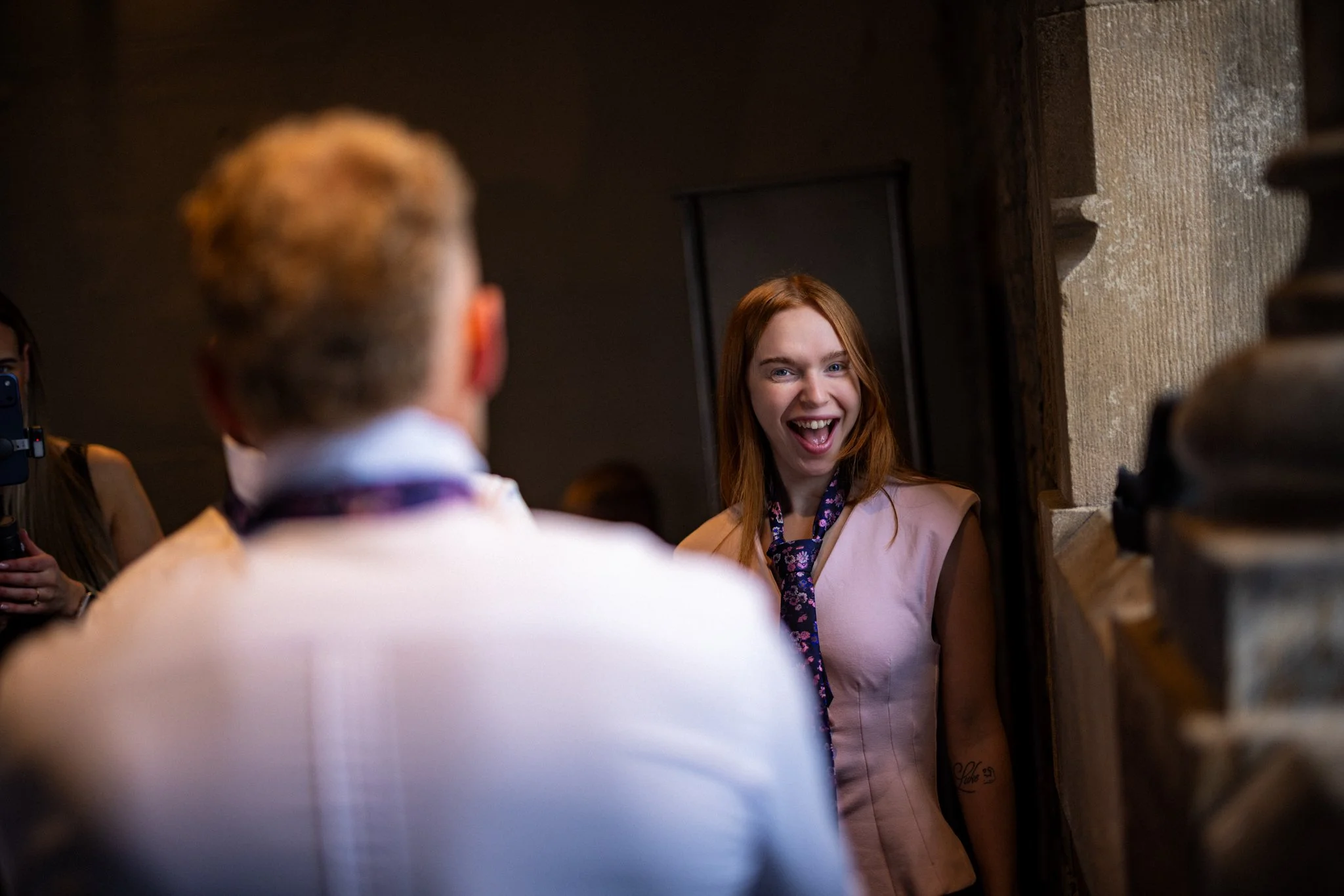 Young woman with red hair smiling and looking at an older person with short light-colored hair, inside a building with stone walls.