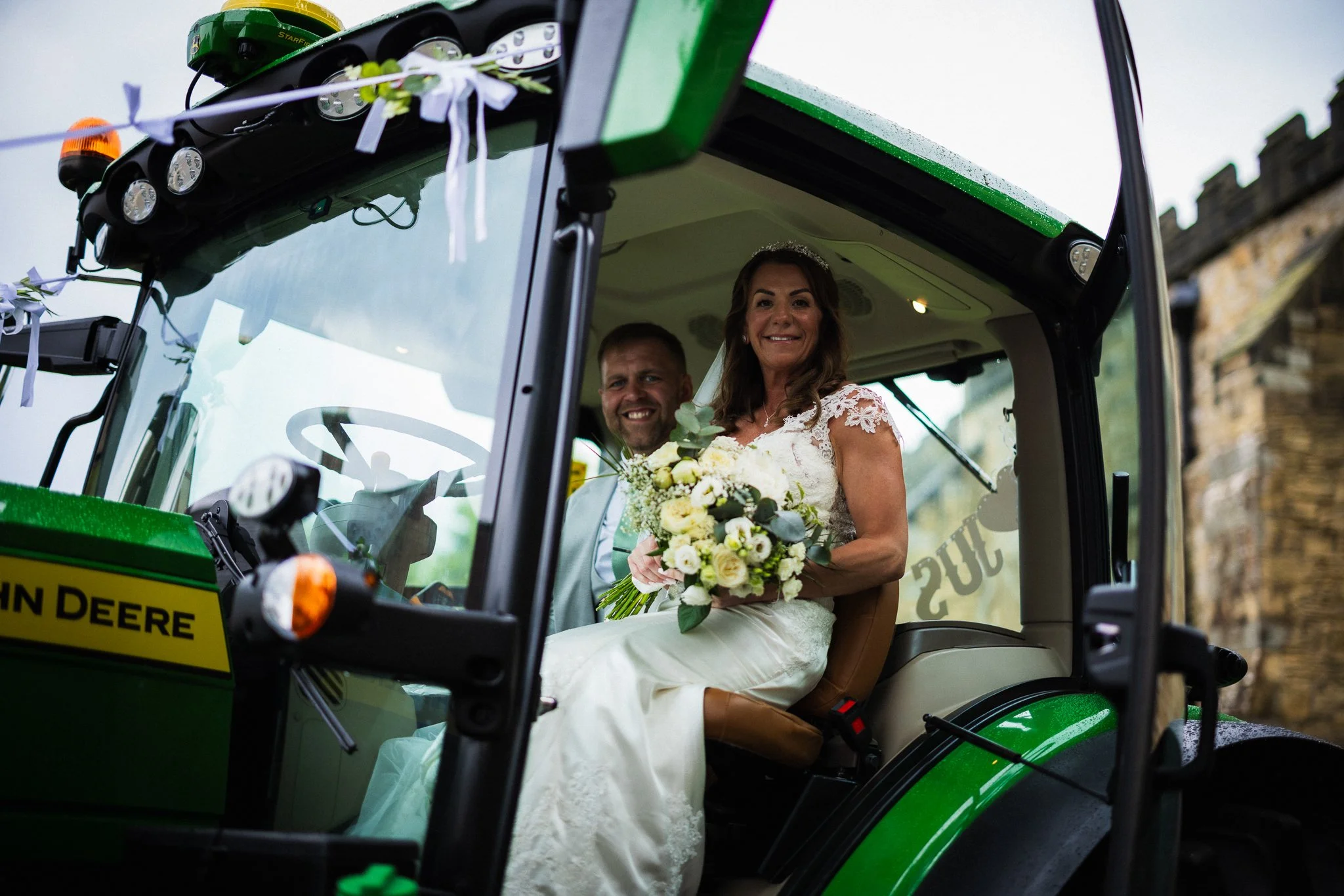 A bride in a wedding dress and a groom smiling inside a green John Deere tractor. The bride is holding a bouquet of white flowers and greenery. The tractor is decorated with ribbons and has a yellow warning sign.