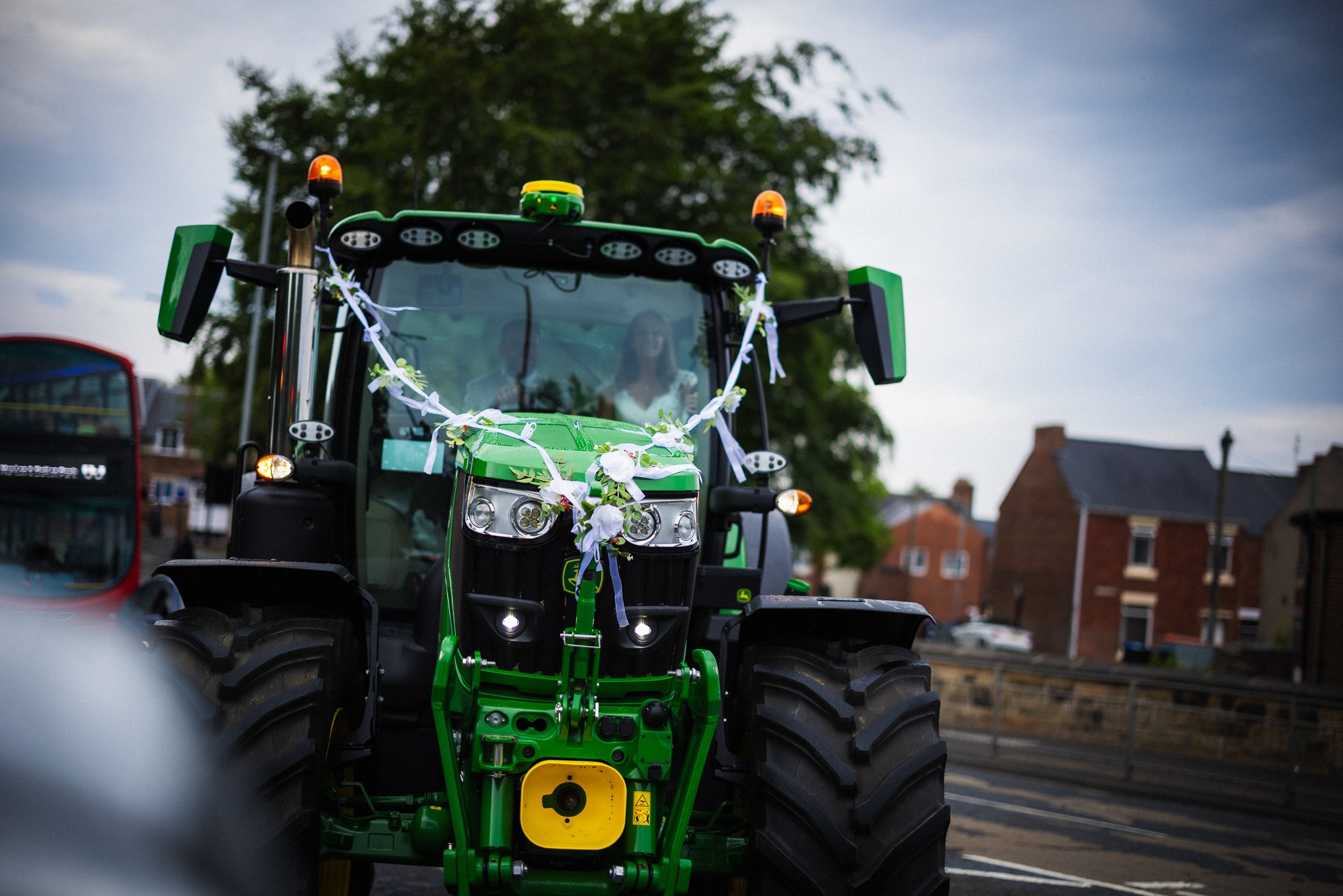 A green tractor decorated with white ribbons and flowers, driven on a street in a residential area with brick houses and a cloudy sky overhead.