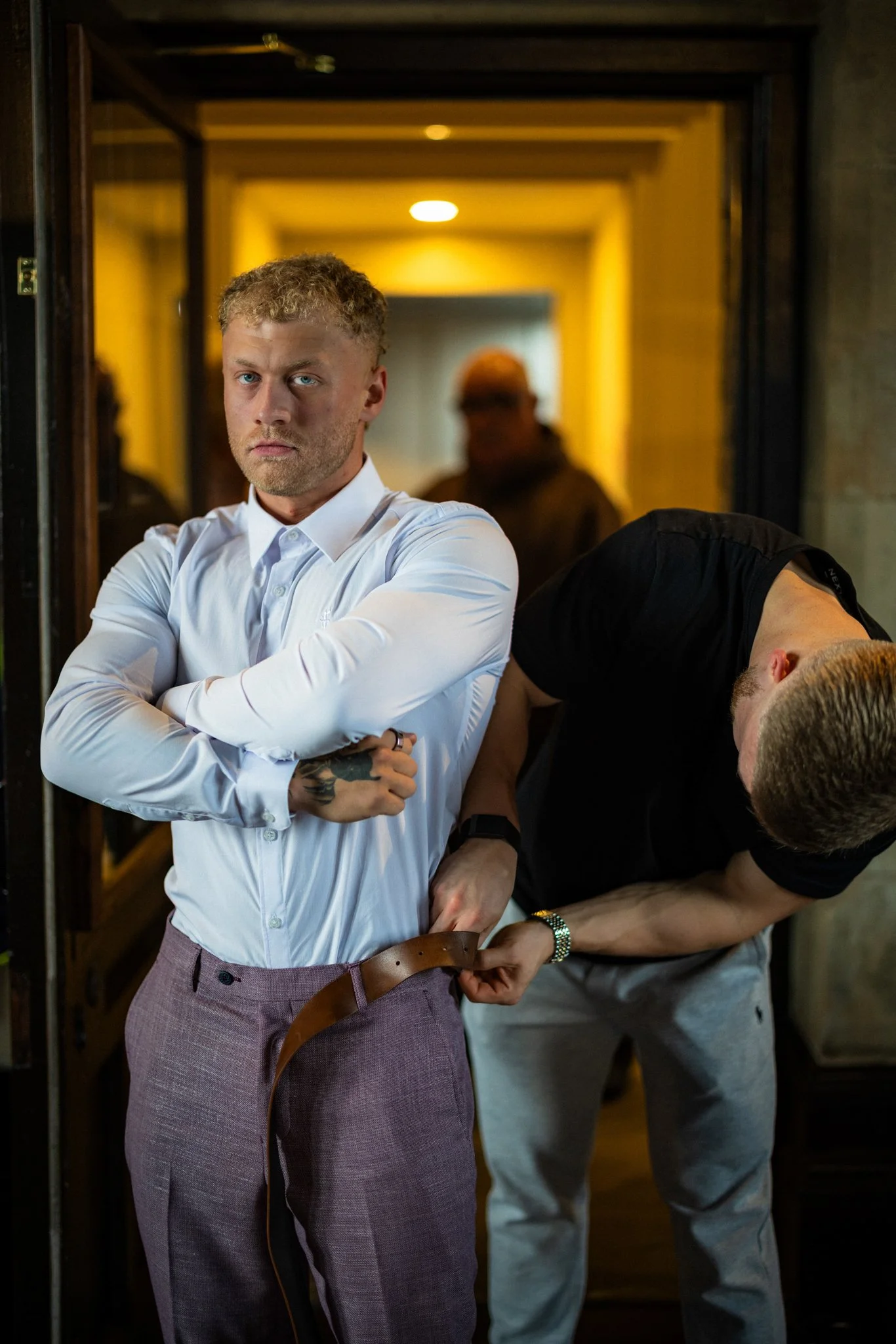A man with light skin, blonde hair, and blue eyes, wearing a white dress shirt and pinkish pants, stands with his arms crossed in a room with warm lighting. Behind him, a man with light skin and reddish hair is helping him adjust a brown belt. A mirr