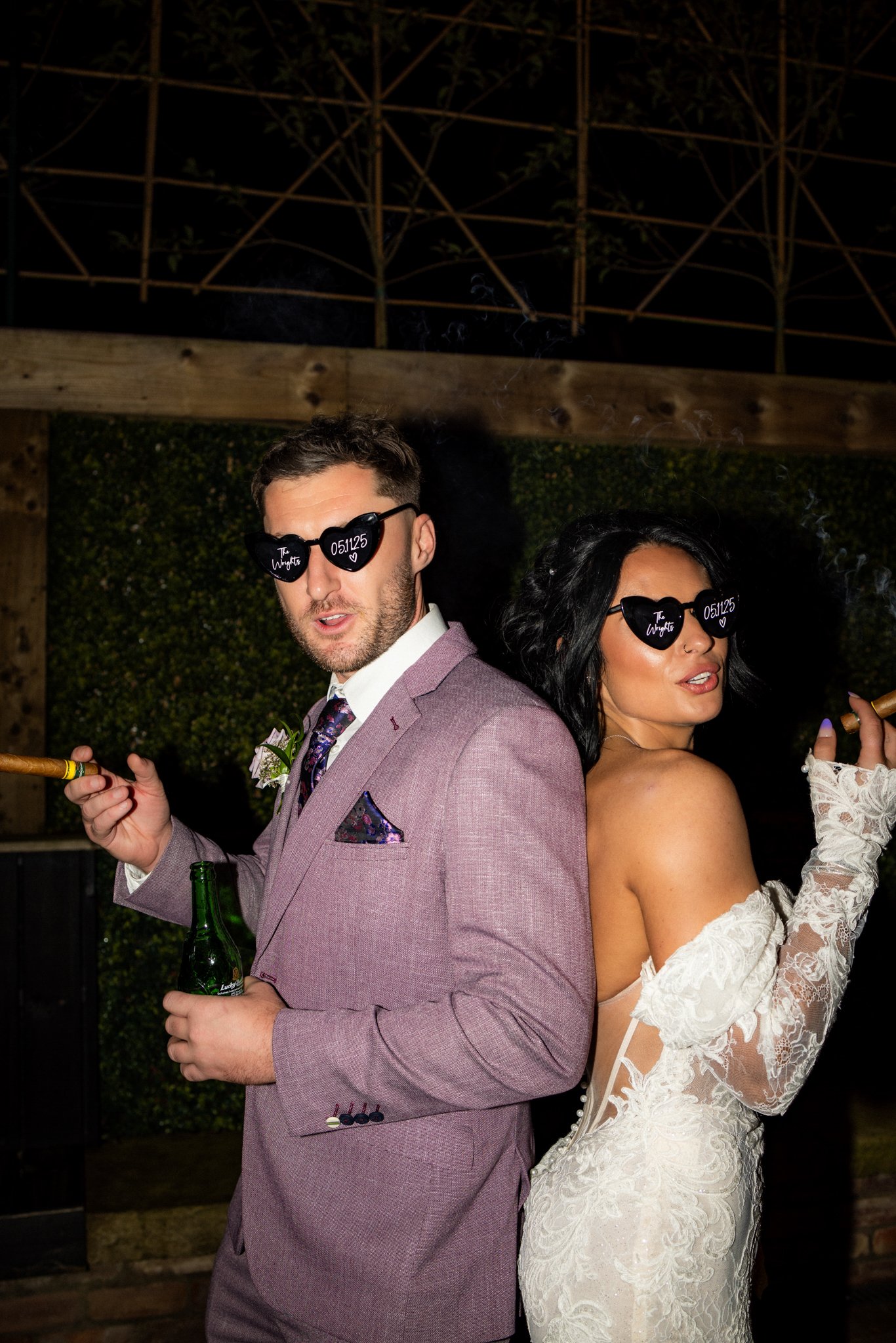 Two people dressed in wedding attire pose back-to-back at night. They are wearing black heart-shaped sunglasses with white writing, holding drinks, and standing outdoors with a wooden fence and greenery in the background.