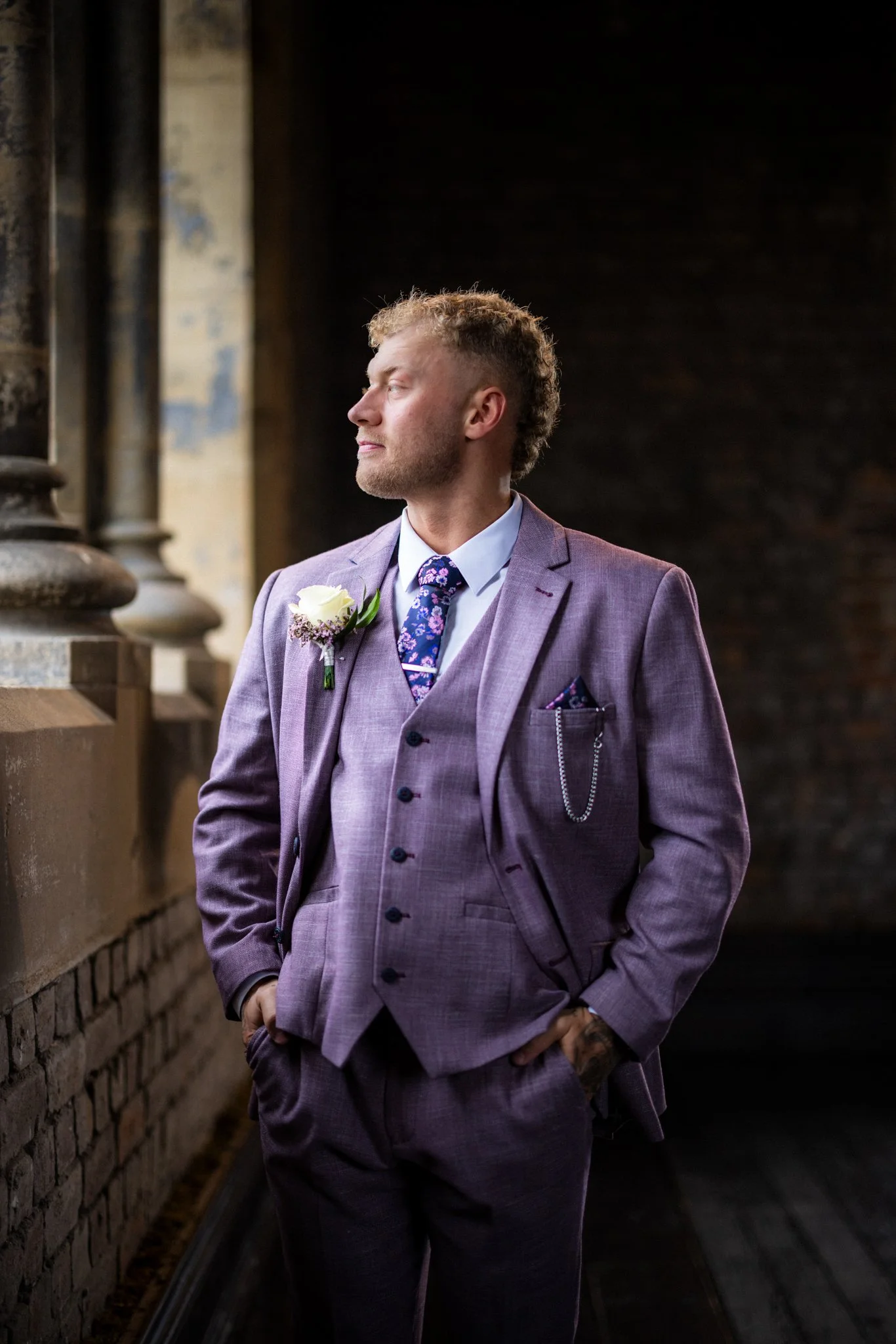 A man with short, curly hair and a beard standing on a dark, narrow walkway with a brick wall and metal pillars on his left, wearing a purple suit, white shirt, and patterned tie, with a white flower boutonniere on his lapel and a chain pocket watch 