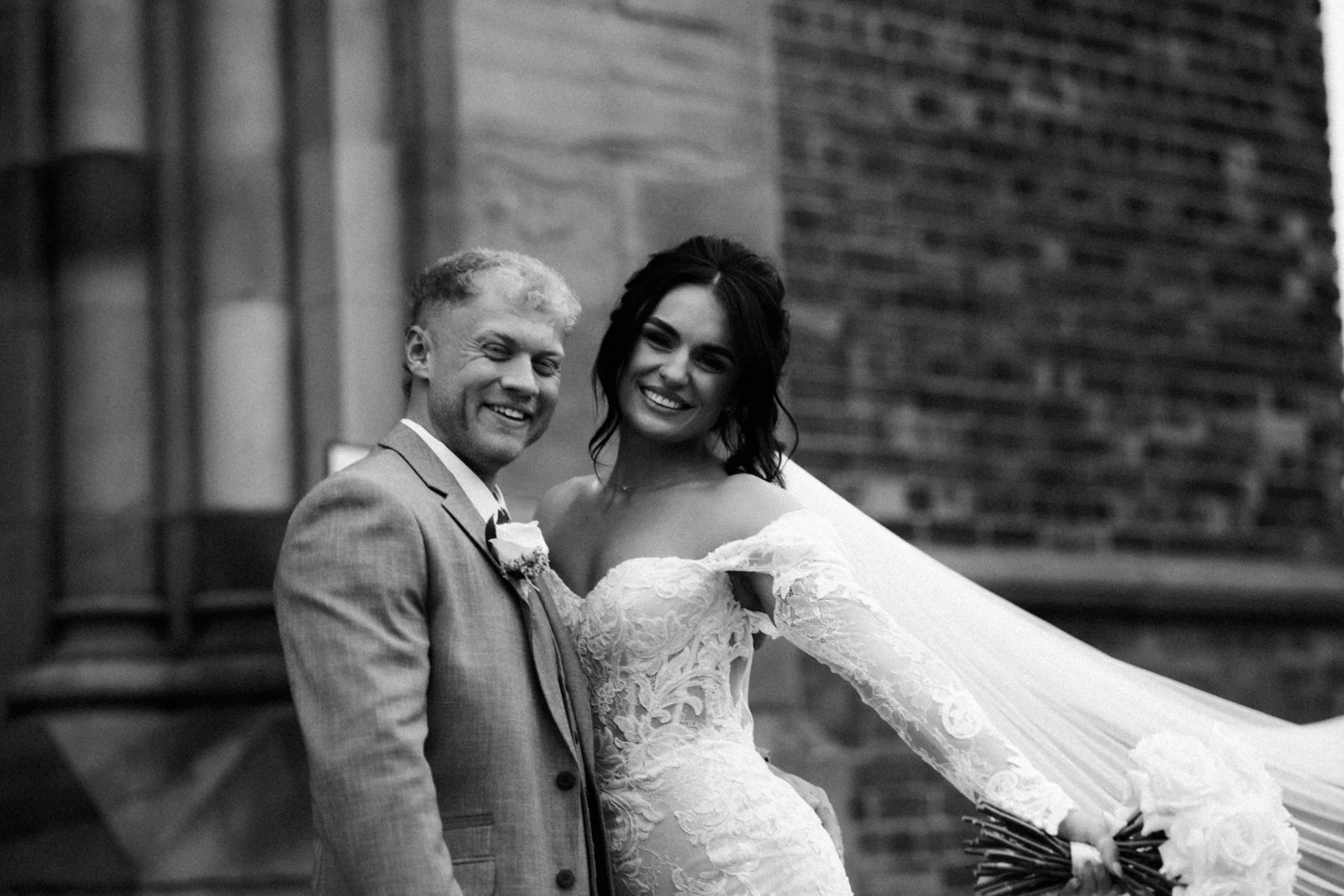 A black and white photo of a smiling couple, the man in a suit and the woman in a lace wedding dress, posing outdoors with a brick wall in the background.