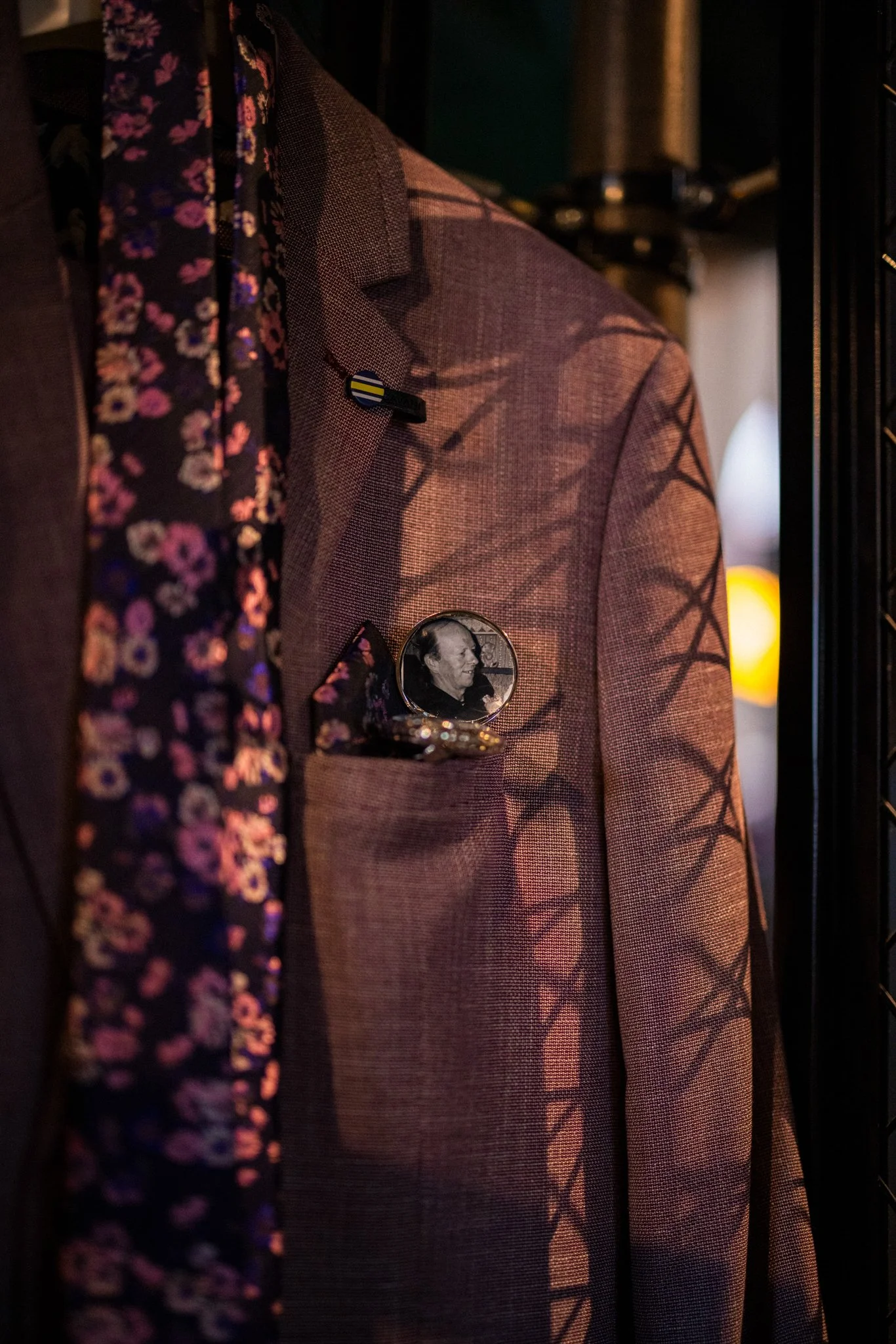 A brown checkered suit jacket with a circular photo button of an elderly man attached to the pocket area, placed next to a floral dress or shirt, in a dimly lit setting.
