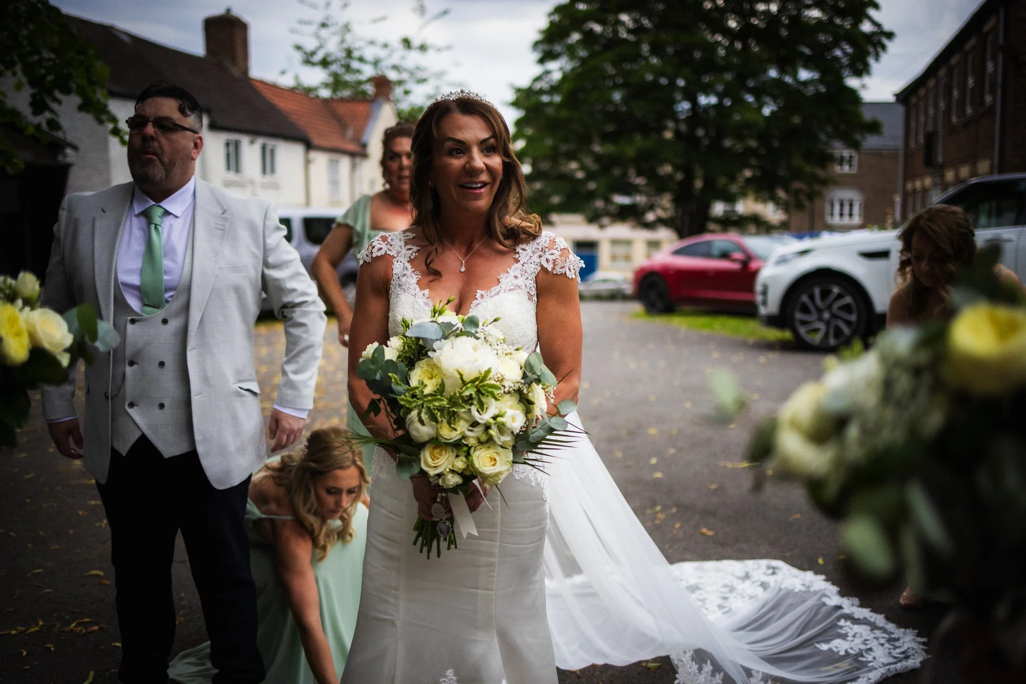 A bride in a white wedding dress holding a bouquet of white roses, standing outdoors surrounded by family or friends, under a tree with cars parked in the background.