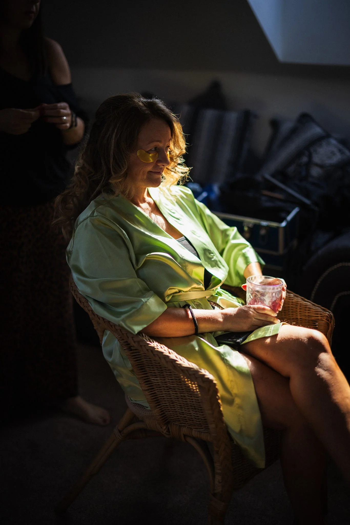 A woman with curly hair wearing yellow eye masks and a shiny green robe sitting in a wicker chair, holding a wine glass, in a dimly lit room.
