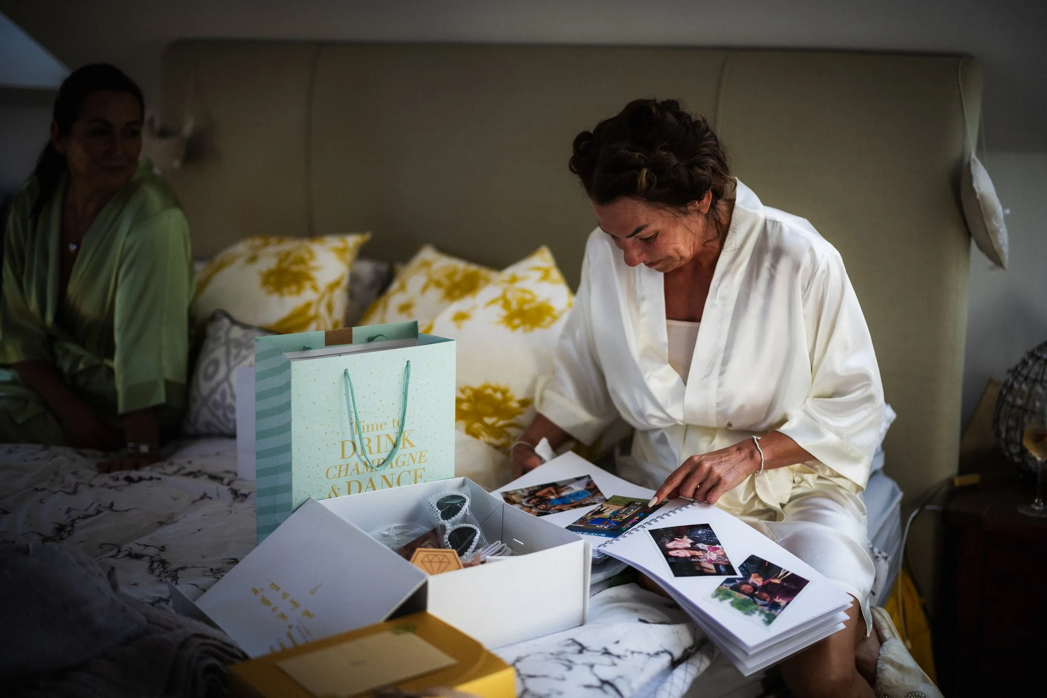 A woman in a white robe sitting on a bed, looking at photo albums and surrounded by photographs, gift bags, and a box of glasses, with another woman partially visible in the background.