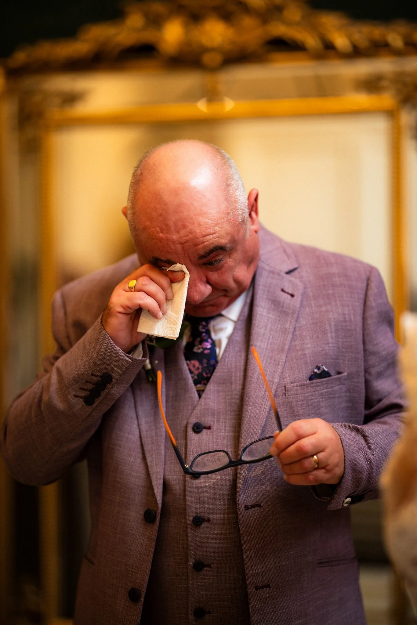 A bald man in a brown plaid suit wipes tears from his eyes with a tissue. He is holding glasses in one hand and has a gold wedding band on his finger.