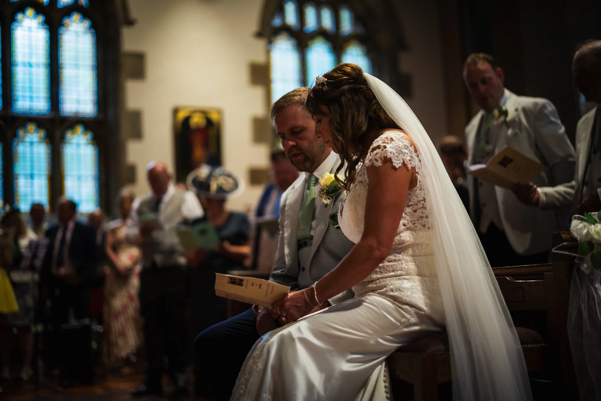 A bride and groom are kneeling in prayer at their wedding ceremony inside a church, surrounded by guests.