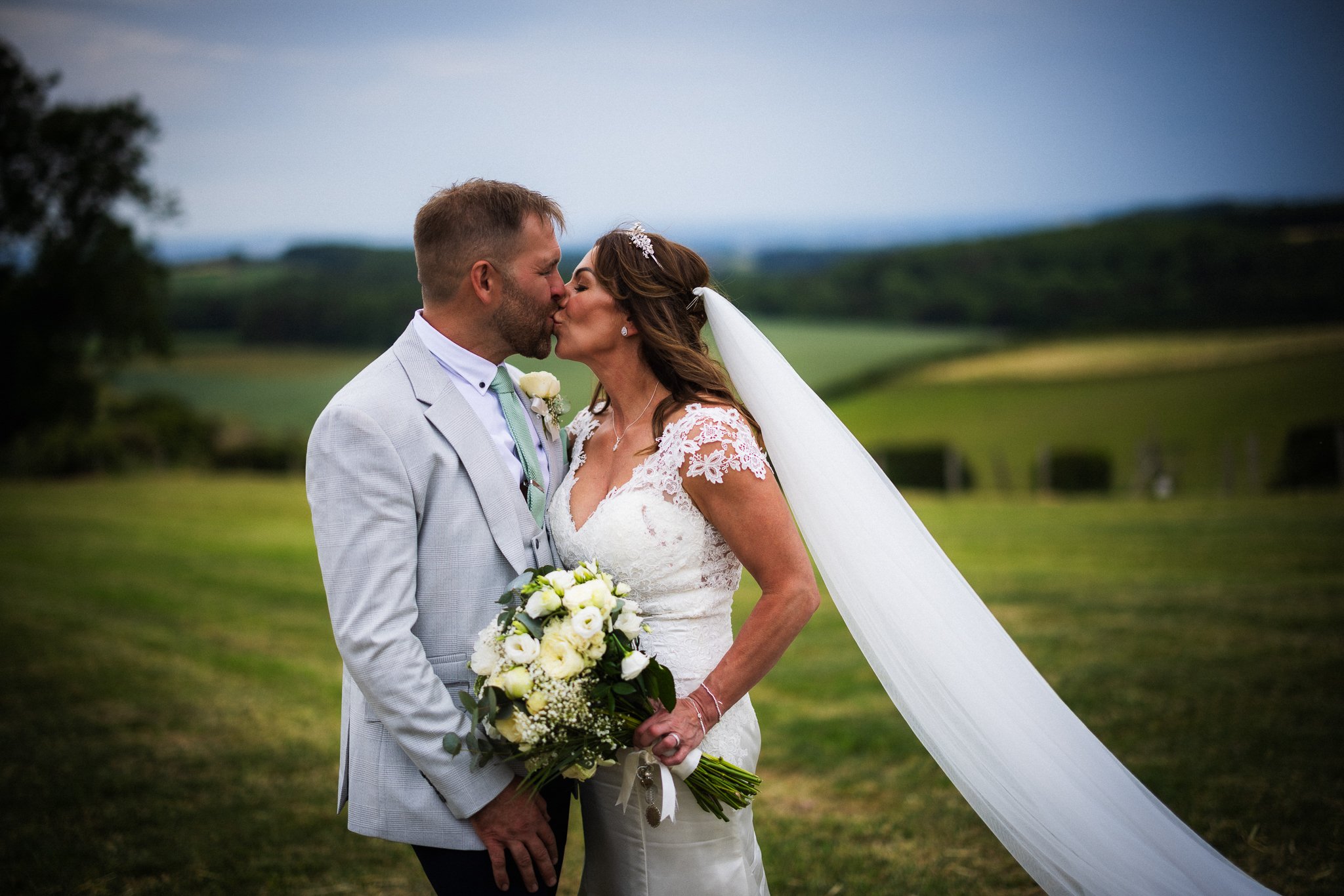 A bride and groom are kissing outdoors during their wedding ceremony, with green fields and cloudy sky in the background. The bride is holding a bouquet of white flowers, and both are dressed in wedding attire.