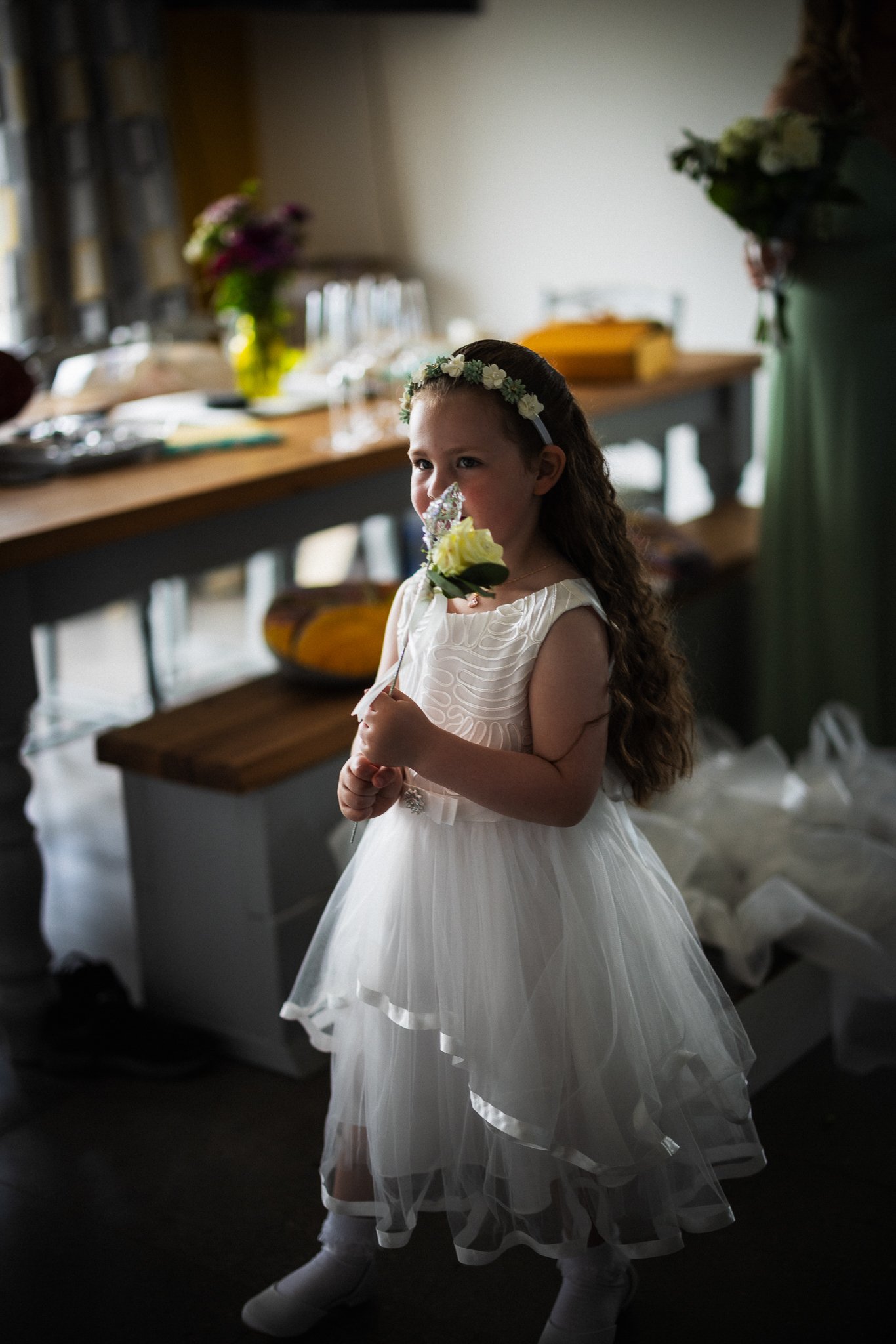 Young girl in a white dress with a floral headband holding a flower, standing indoors near a table with flower arrangements and glasses.