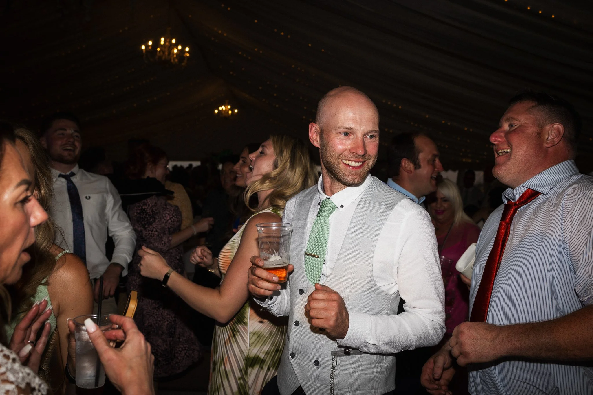People in formal attire socializing at a party or wedding reception inside a decorated tent with chandeliers.