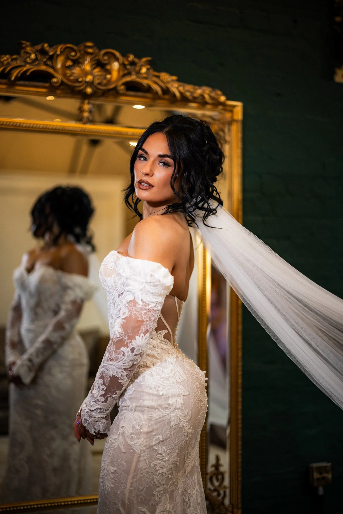 A woman in a white lace wedding dress with long sleeves and an off-the-shoulder design, standing in front of a large ornate gold-framed mirror, with her reflection visible. She has dark hair styled in loose curls and is looking at the camera with a c