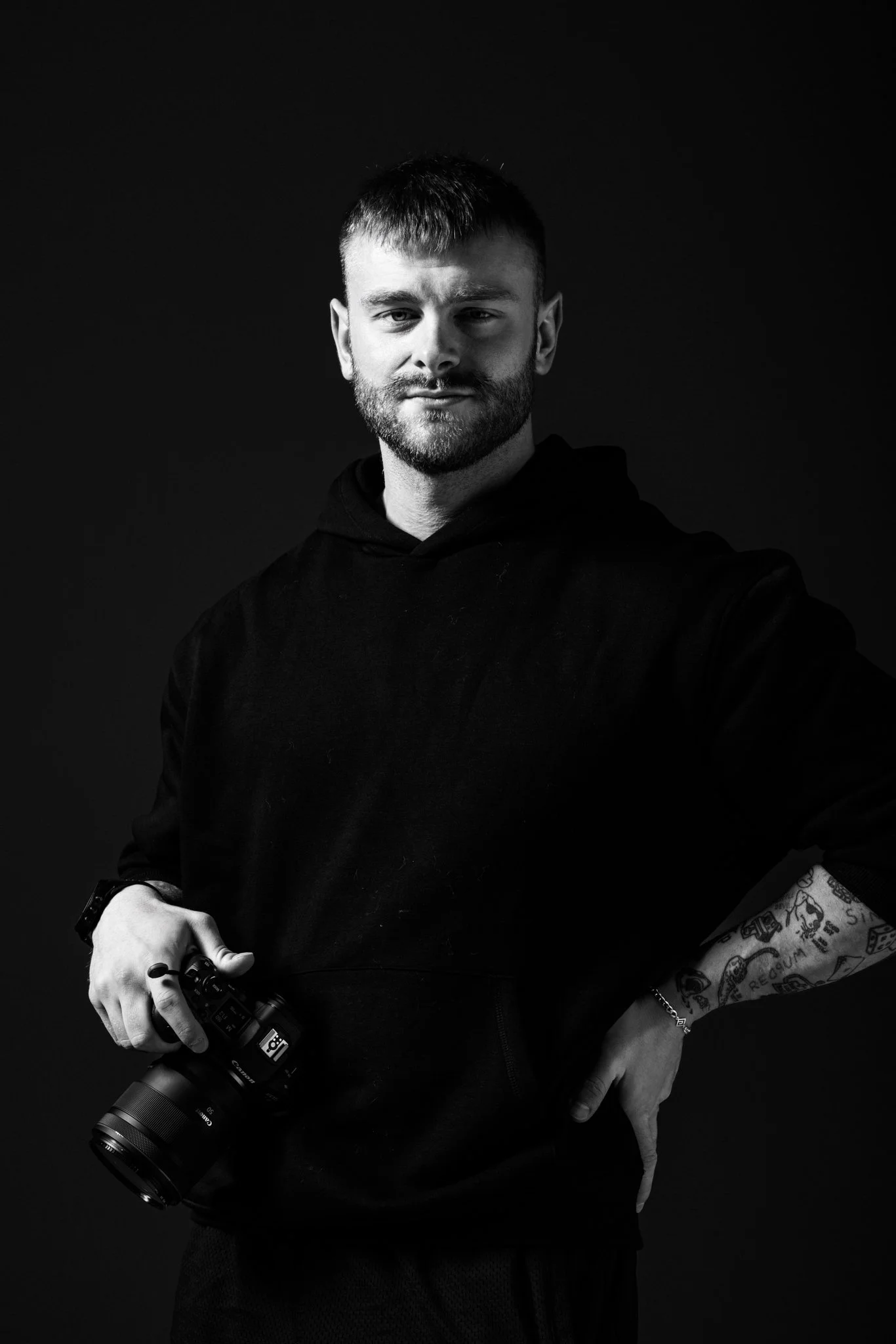 Black and white portrait of a young man with short hair and beard, wearing a black hoodie, holding a professional camera, with part of his tattooed arm visible, against a dark background.