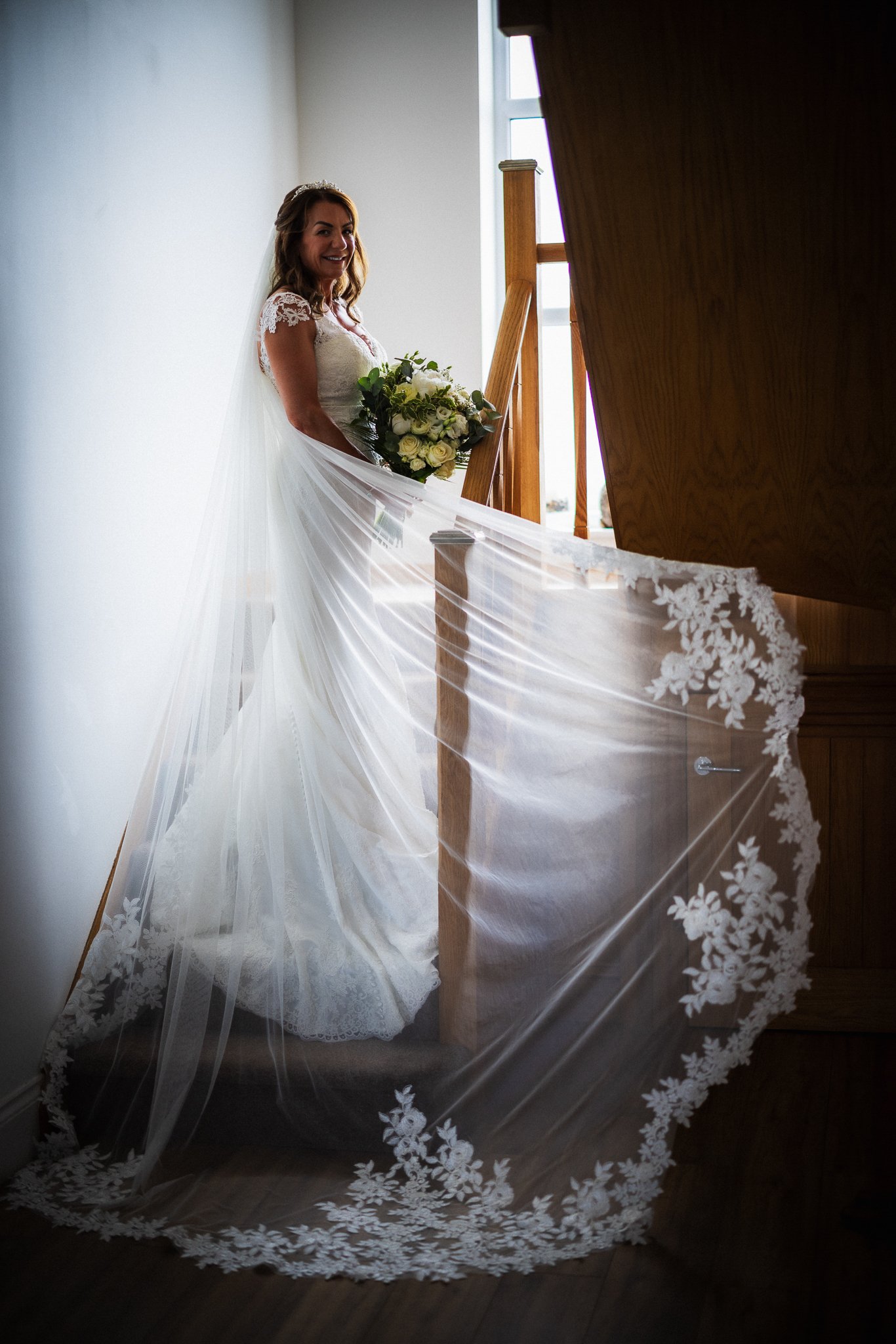 A smiling bride in a white wedding dress holding a bouquet, standing next to a piano with a sheer lace-embellished veil cascading from her dress.