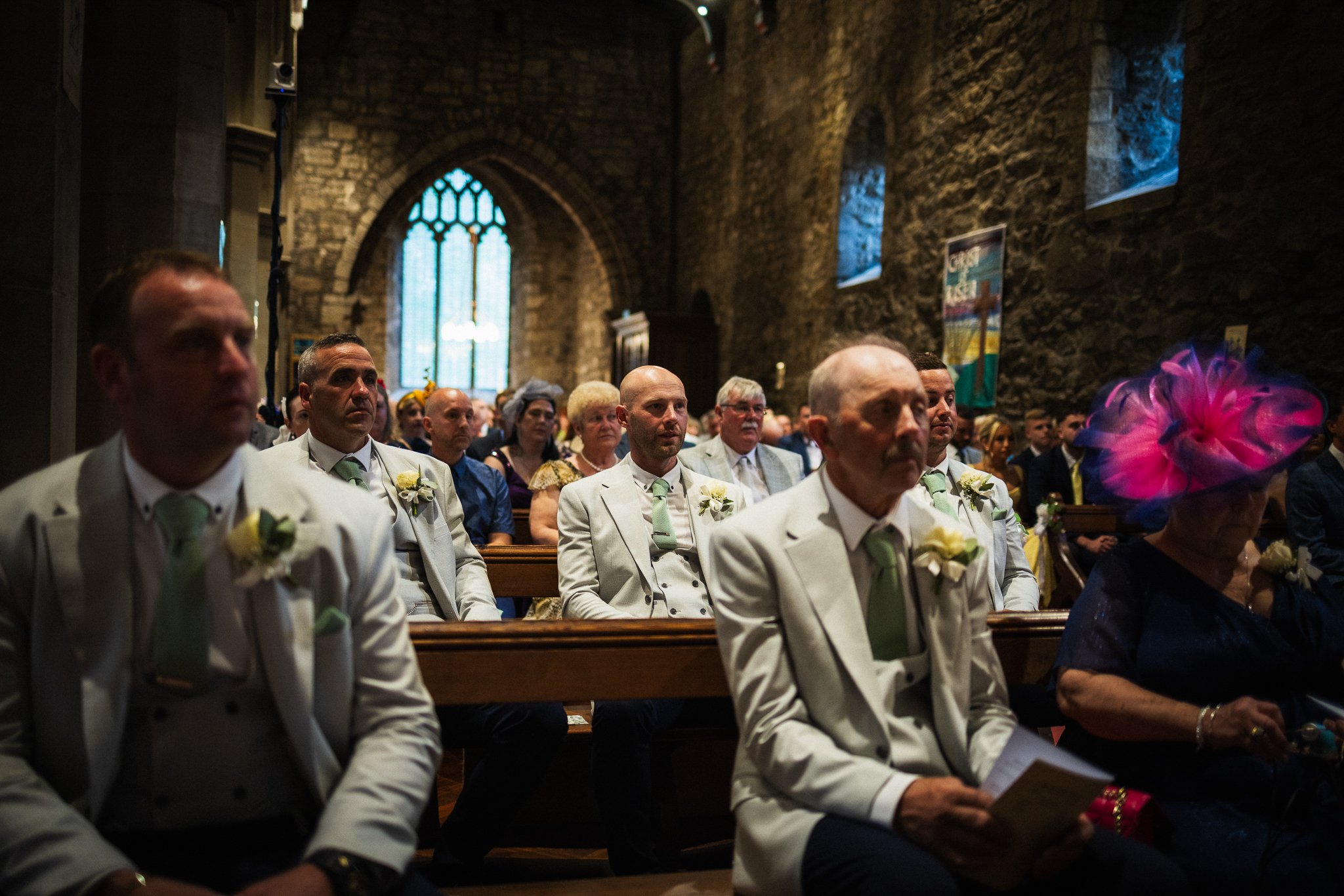 A group of wedding guests sitting in pews inside a stone church with stained glass windows, dressed in formal attire, attending a wedding ceremony.