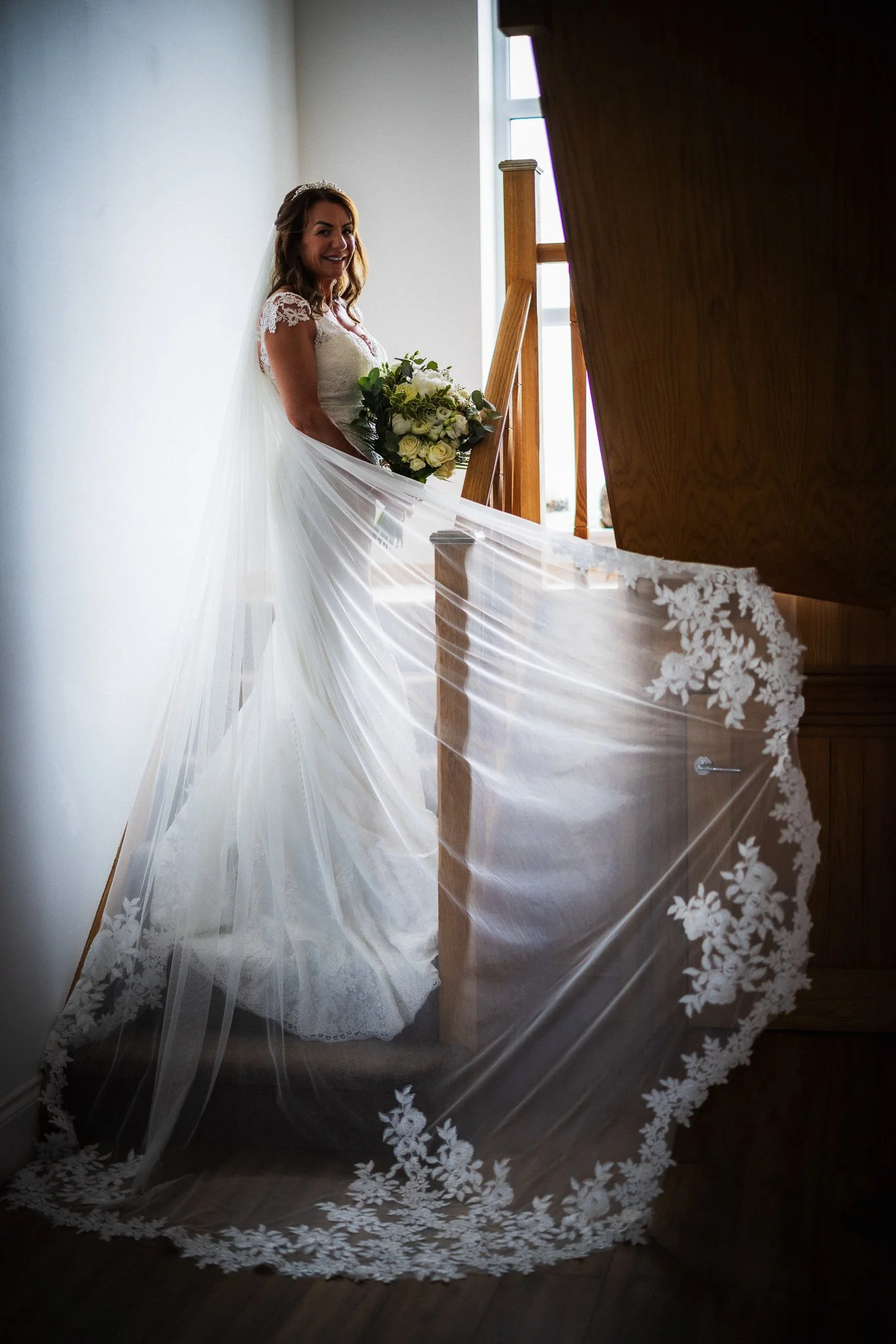 A bride in a white wedding dress holding a bouquet of white flowers, standing indoors near a staircase with a window in the background.