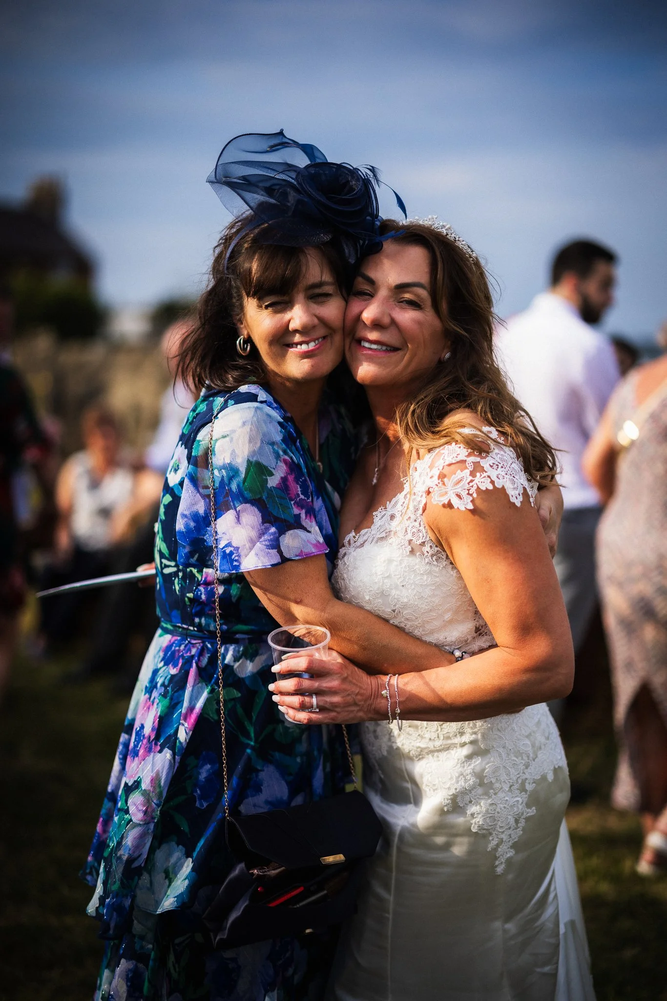 Two women hugging at an outdoor event during sunset, one in a colorful dress with a large dark blue hat, the other in a white lace dress and tiara, both smiling.