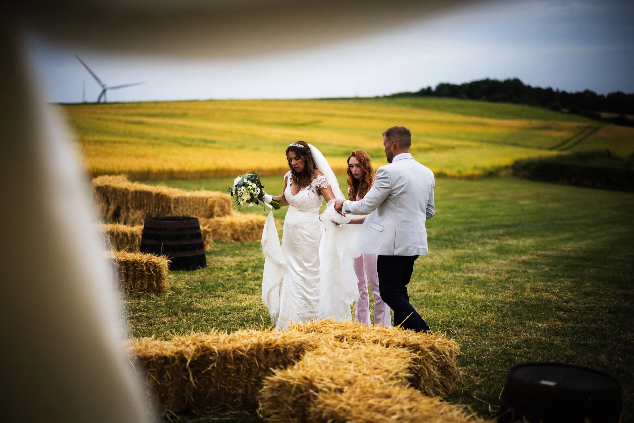 A bride and groom holding hands during their wedding ceremony outdoors in a rural setting, with hay bales and rolling green fields in the background.
