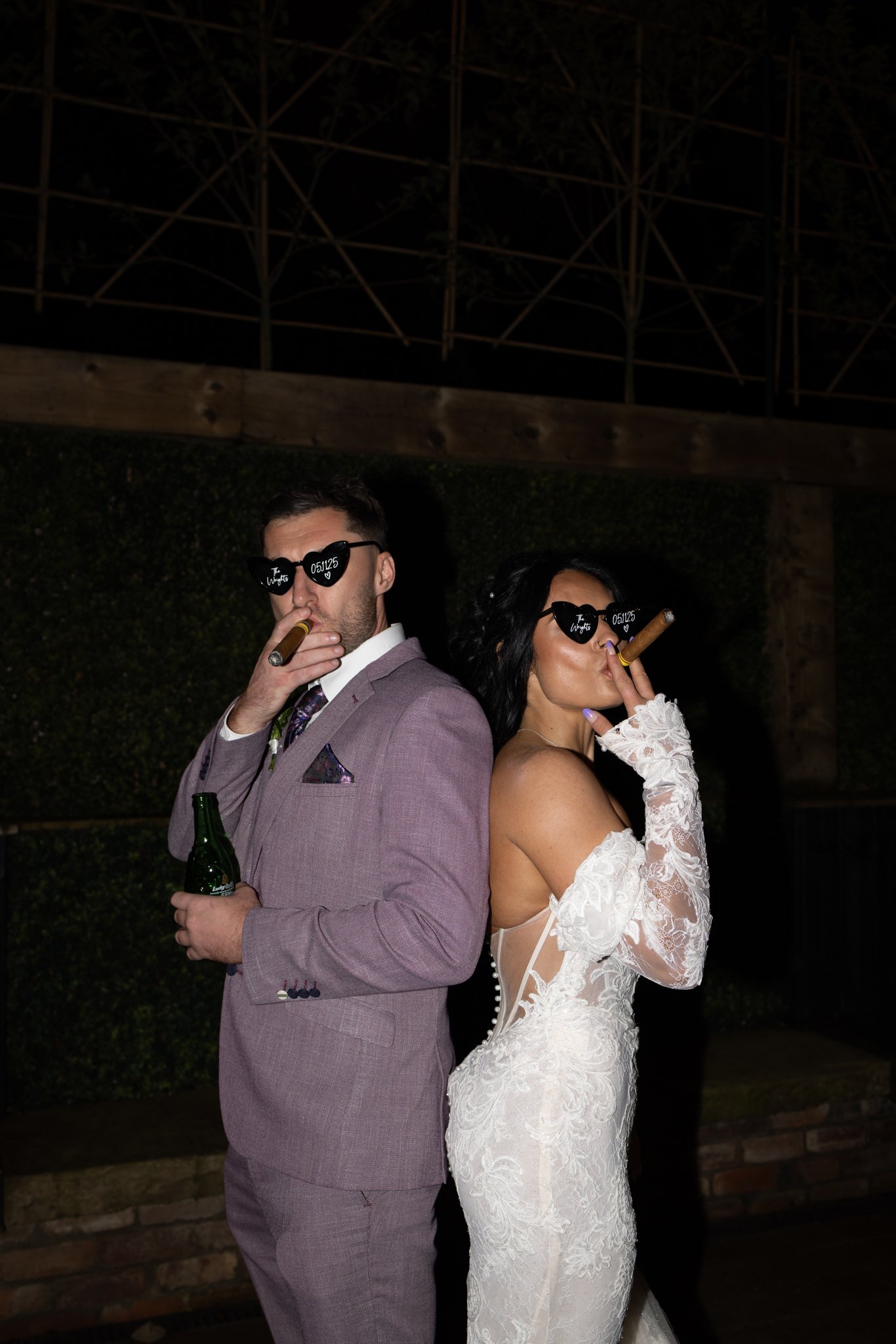 A man in a purple suit jacket and a woman in a white lace dress and gloves, both wearing sunglasses with writing on the lenses, smoking cigars, holding green glass bottles, standing back to back outdoors at night.