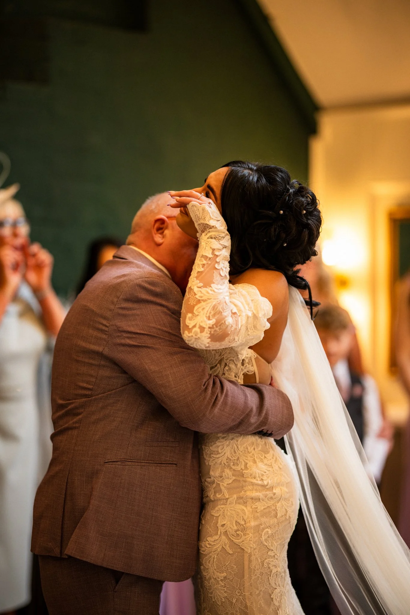 A wedding ceremony with a bride in a lace wedding gown and a groom in a brown suit sharing an emotional moment, possibly a kiss, during the event with guests in the background.