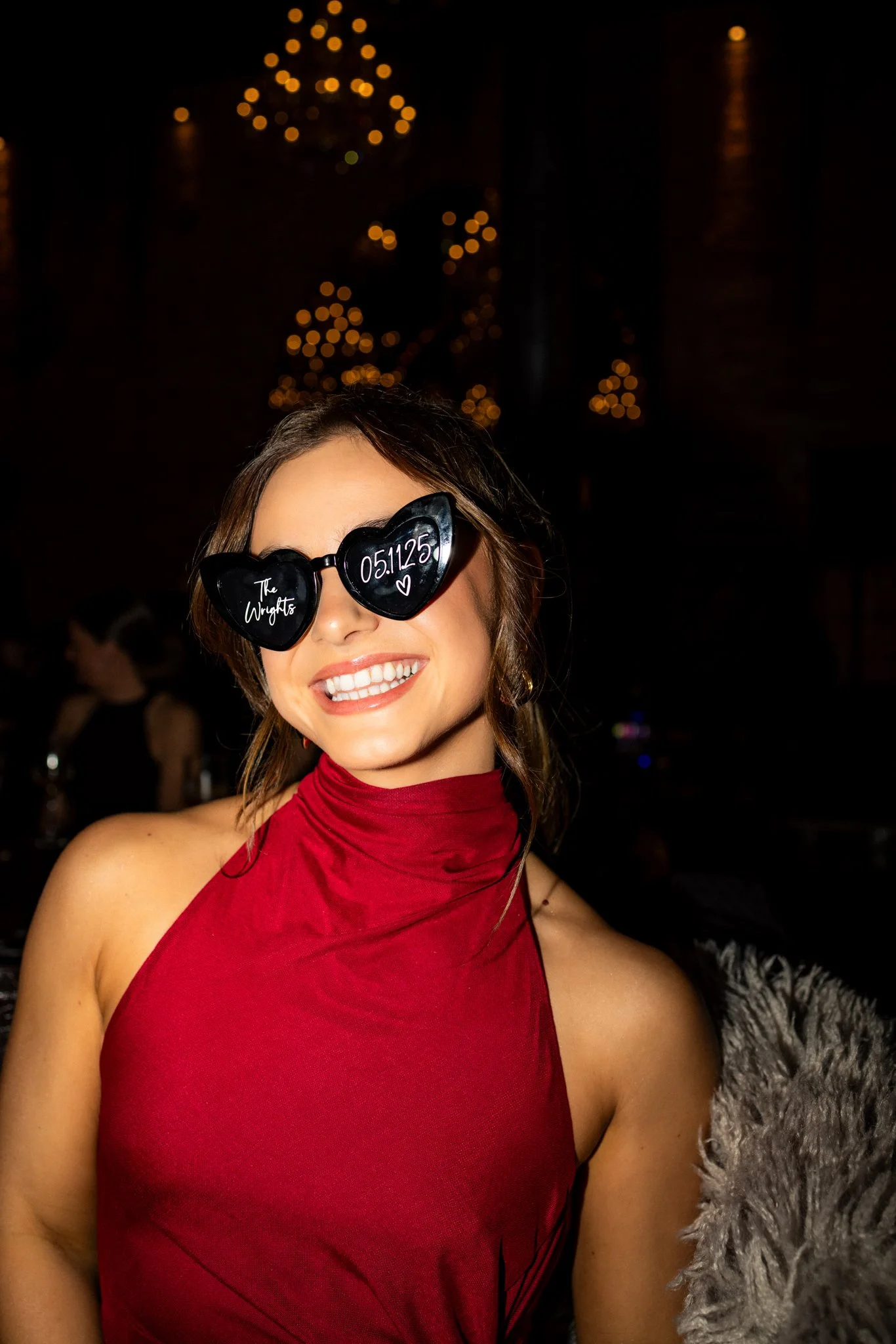 A smiling woman wearing heart-shaped sunglasses and a red sleeveless dress, standing indoors at a celebration or party with blurry lights and people in the background.