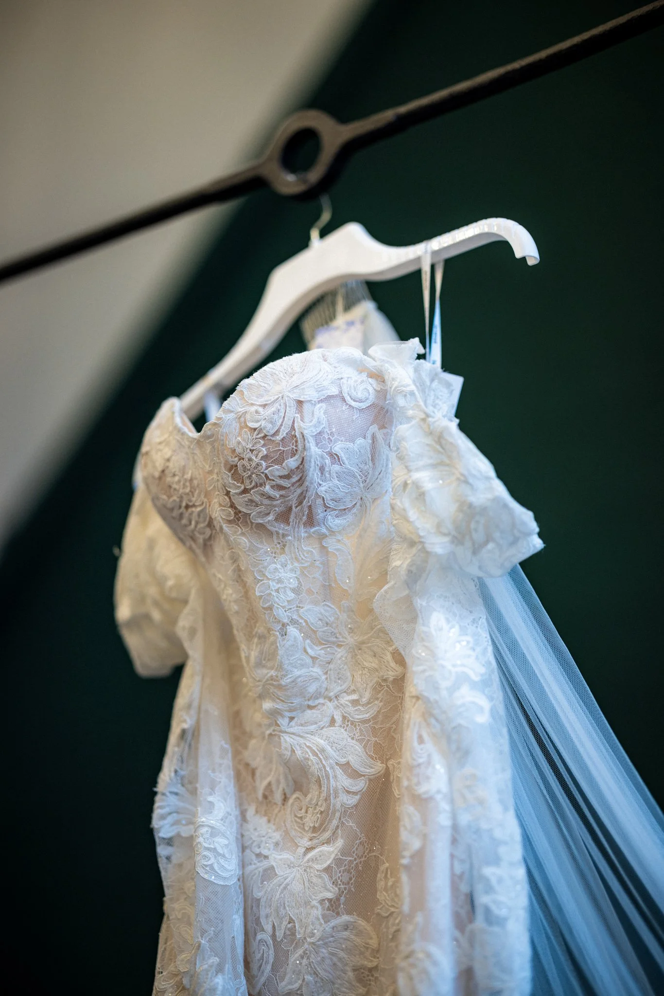 A wedding dress with lace and floral embroidery hanging on a white hanger against a dark background.