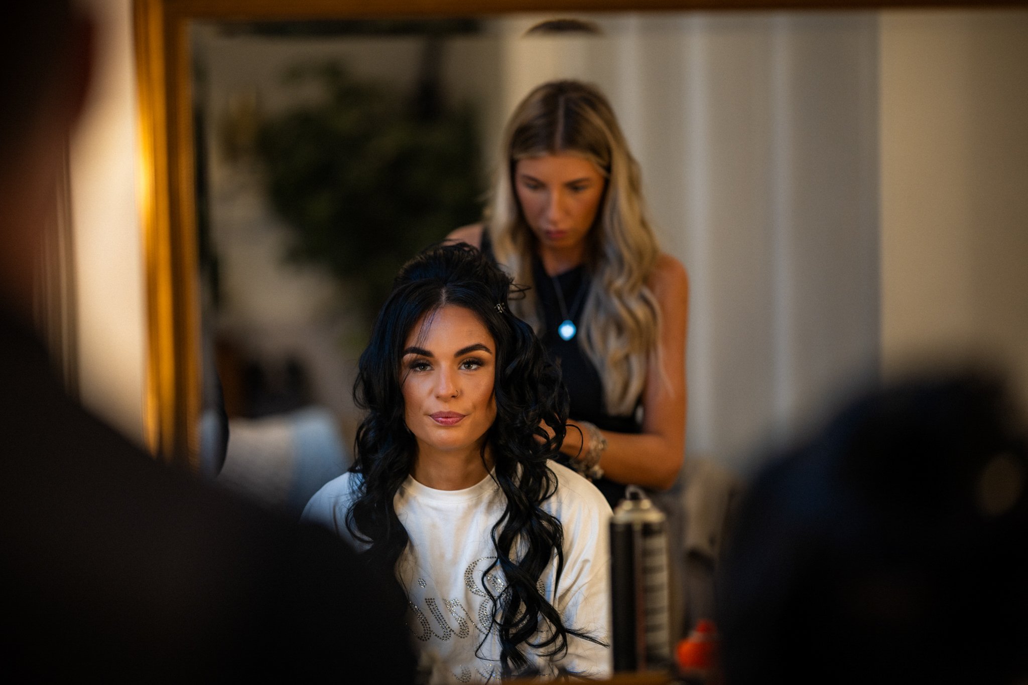 A woman with long black hair sitting in front of a mirror getting her hair styled, with a stylist working behind her. The mirror reflects her face and part of her outfit.
