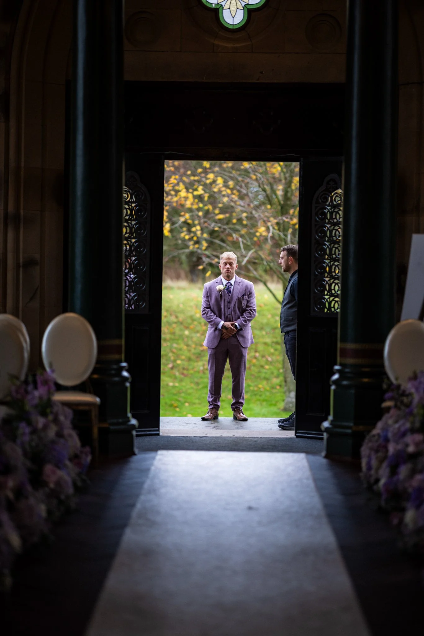 Man in a light purple suit standing outside a church entrance, seen through an open doorway with decorative wooden panels, with trees in the background.