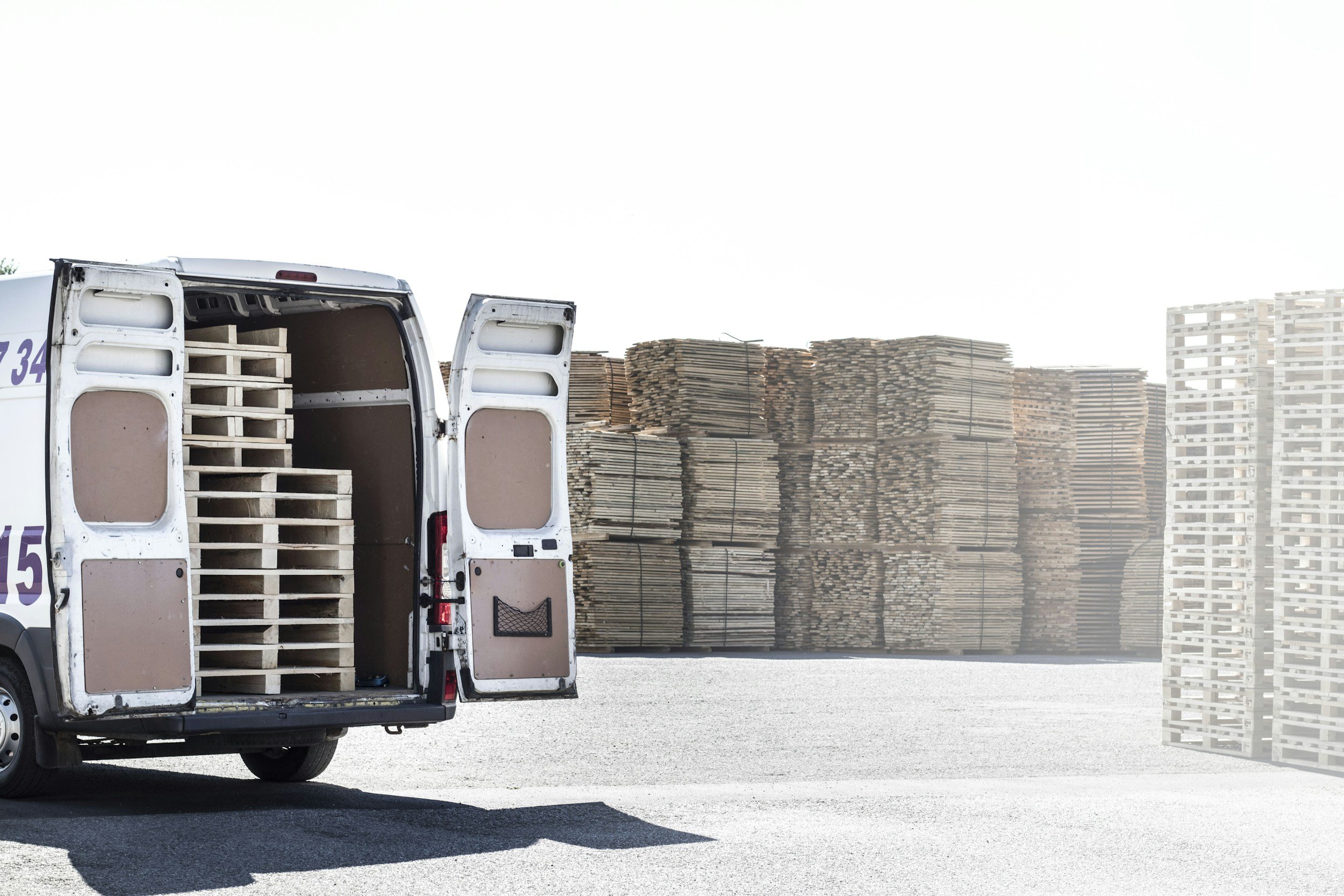 A white delivery van with open rear doors revealing wooden pallets inside, parked on an asphalt lot with large stacks of cardboard boxes or pallets in the background.