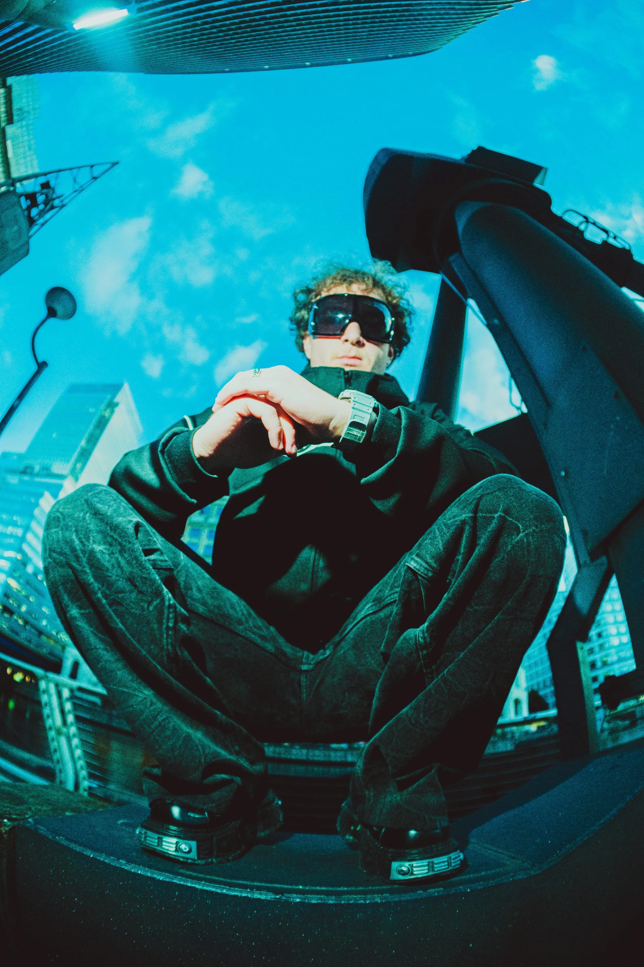 A person with curly hair wearing sunglasses and black clothing taking a selfie in a mirror on a city street, with tall buildings and a blue sky in the background.