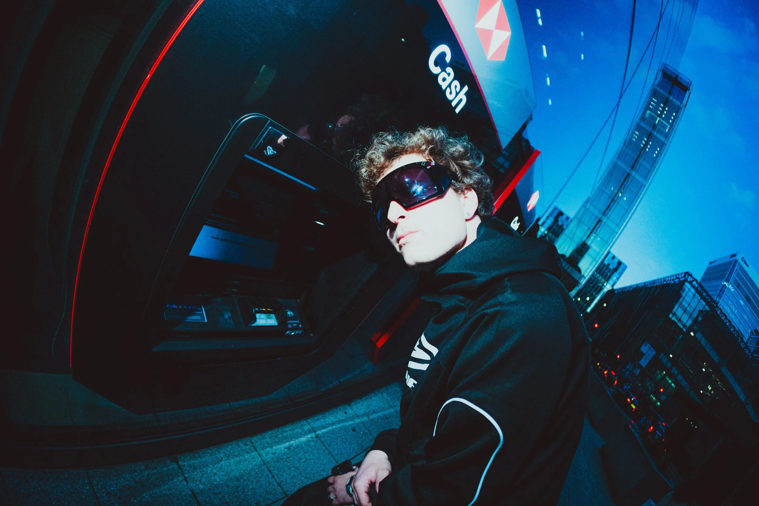 A person with curly hair wearing large sunglasses, a black jacket, and a black hoodie, stands in front of a vending machine at dusk in an urban environment with tall glass buildings and blue sky. The vending machine has a sign that reads 'Cash' and images of snacks.